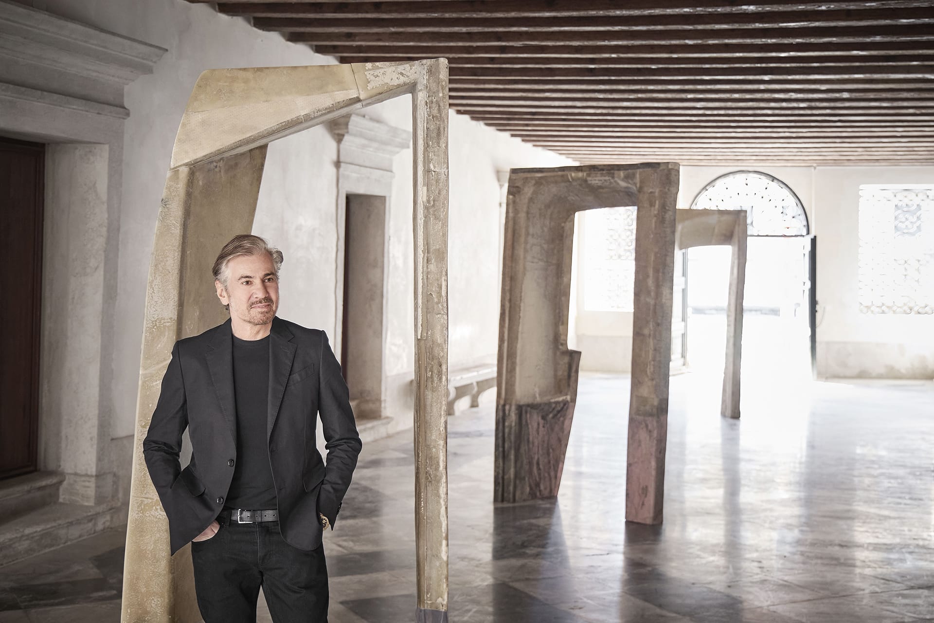 A person in a black suit standing in an art gallery with abstract sculptures in a bright, spacious room.