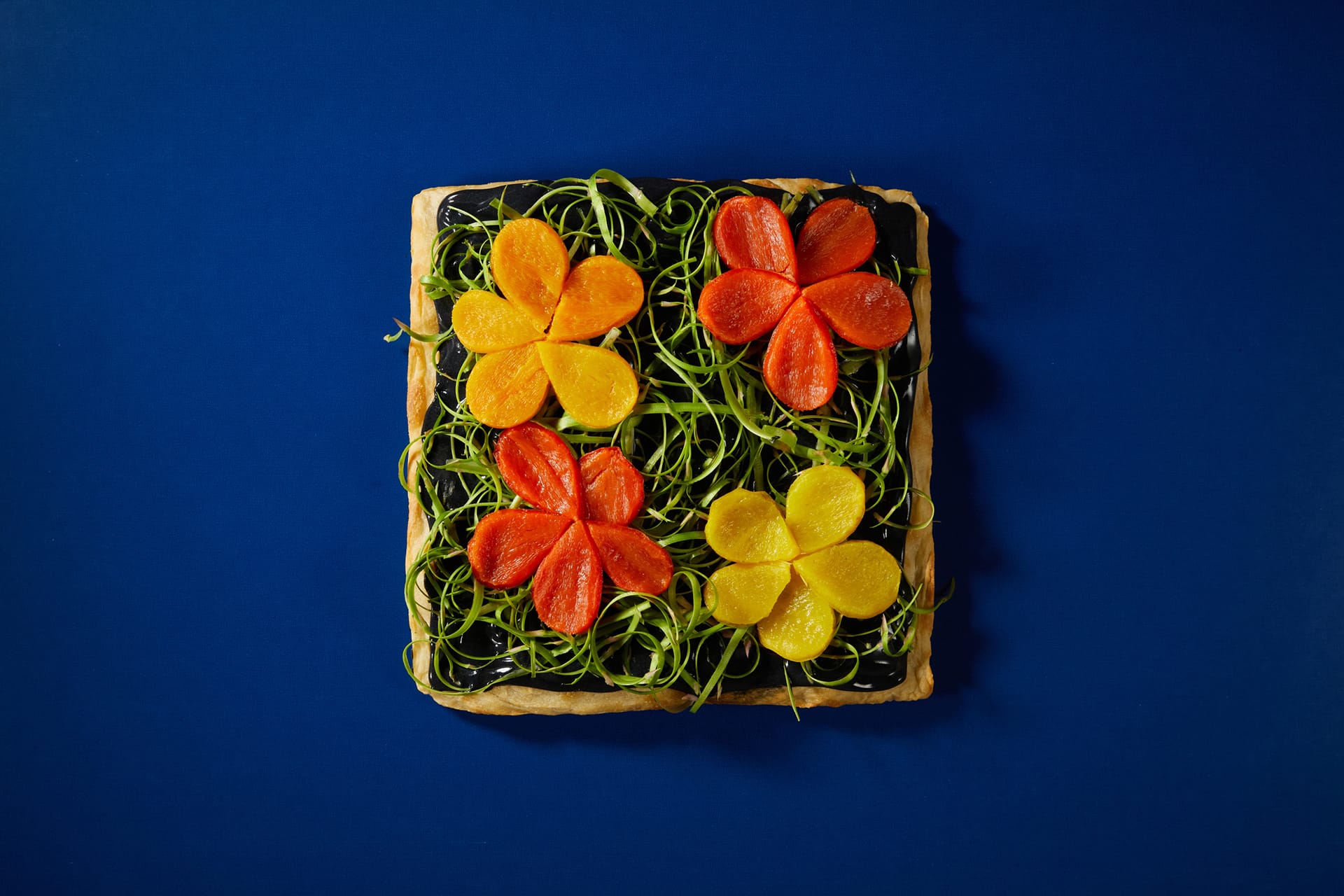 Square tart with vibrant orange and yellow flower-shaped toppings on a bed of green, set against a deep blue background.