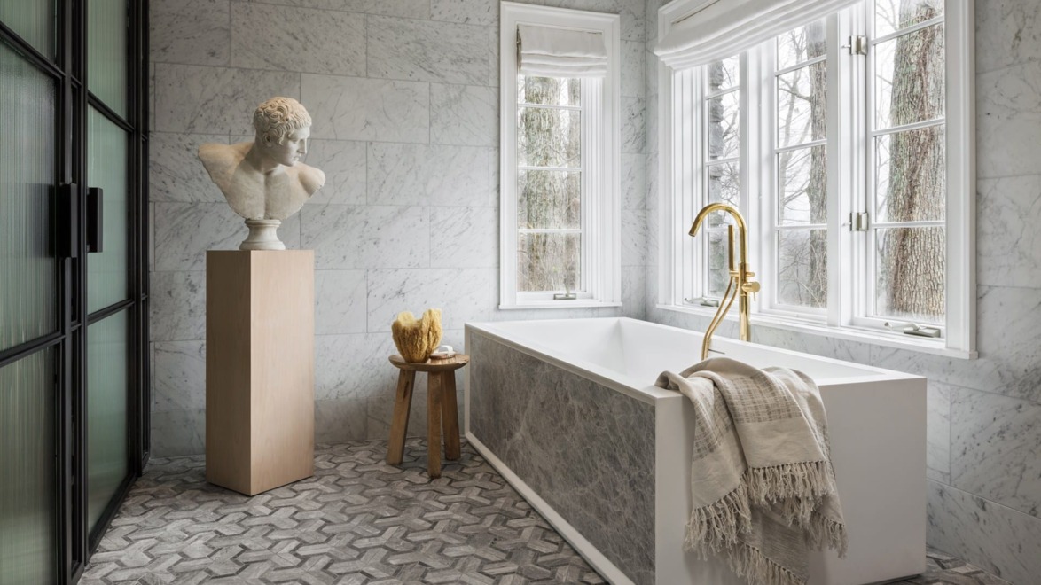 Luxurious bathroom with marble walls, freestanding tub, gold faucet, bust sculpture on pedestal, and large window.