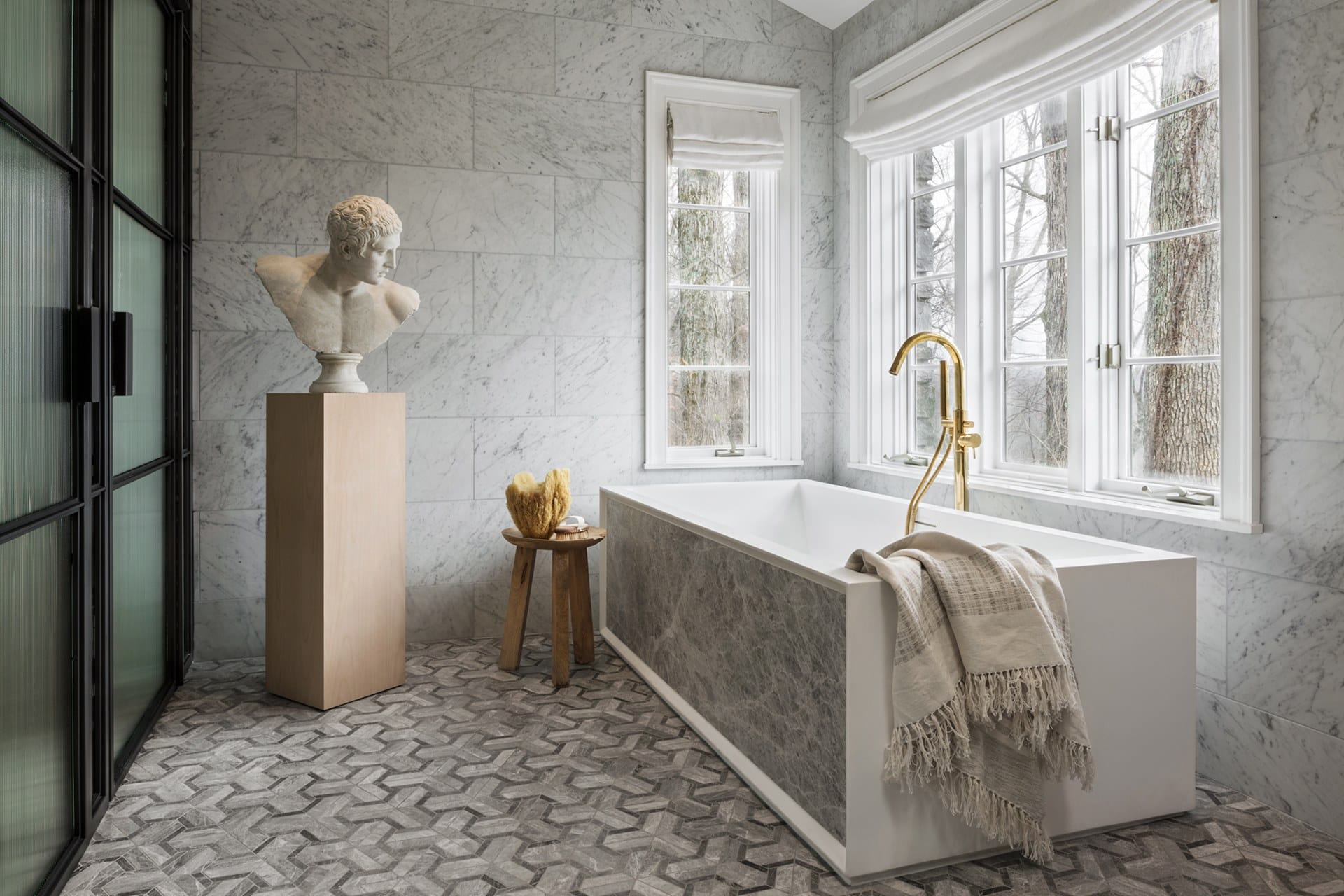 Luxurious bathroom with marble walls, freestanding tub, gold faucet, bust sculpture on pedestal, and large window.