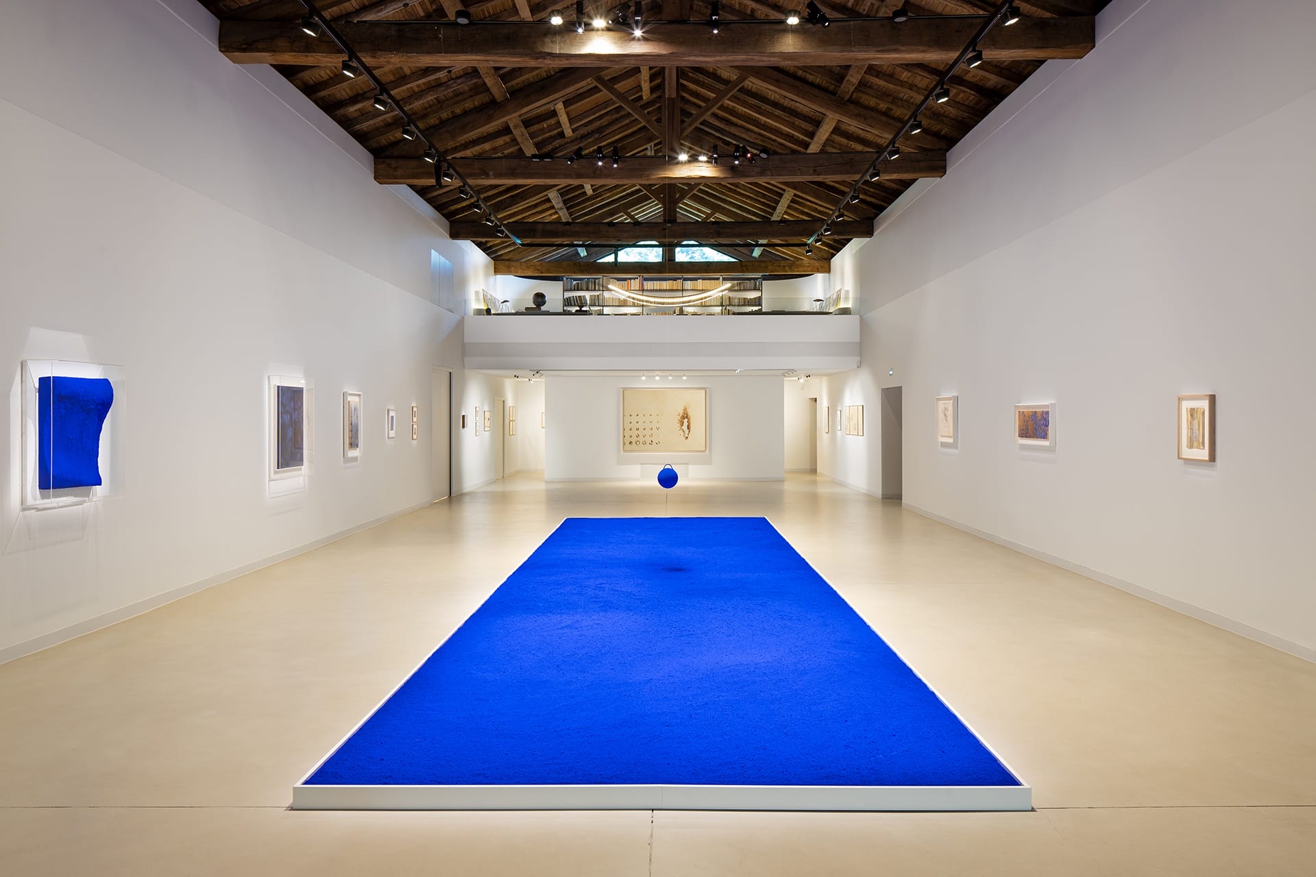 Spacious art gallery with wooden ceiling, blue carpet centerpiece, and framed artworks on white walls.