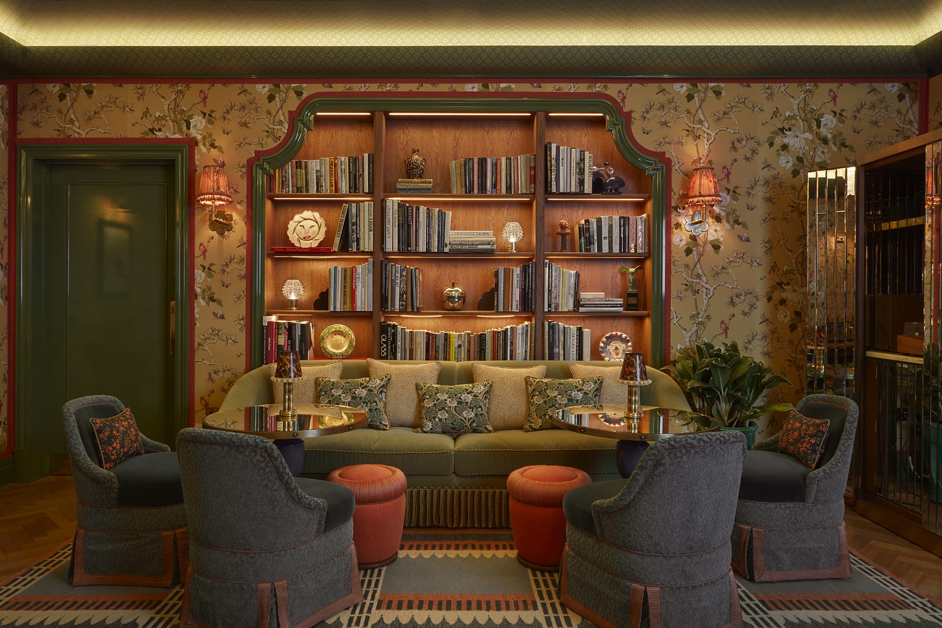 Stylish vintage living room with floral wallpaper, elegant bookshelf, green sofa, round tables, and plush chairs.