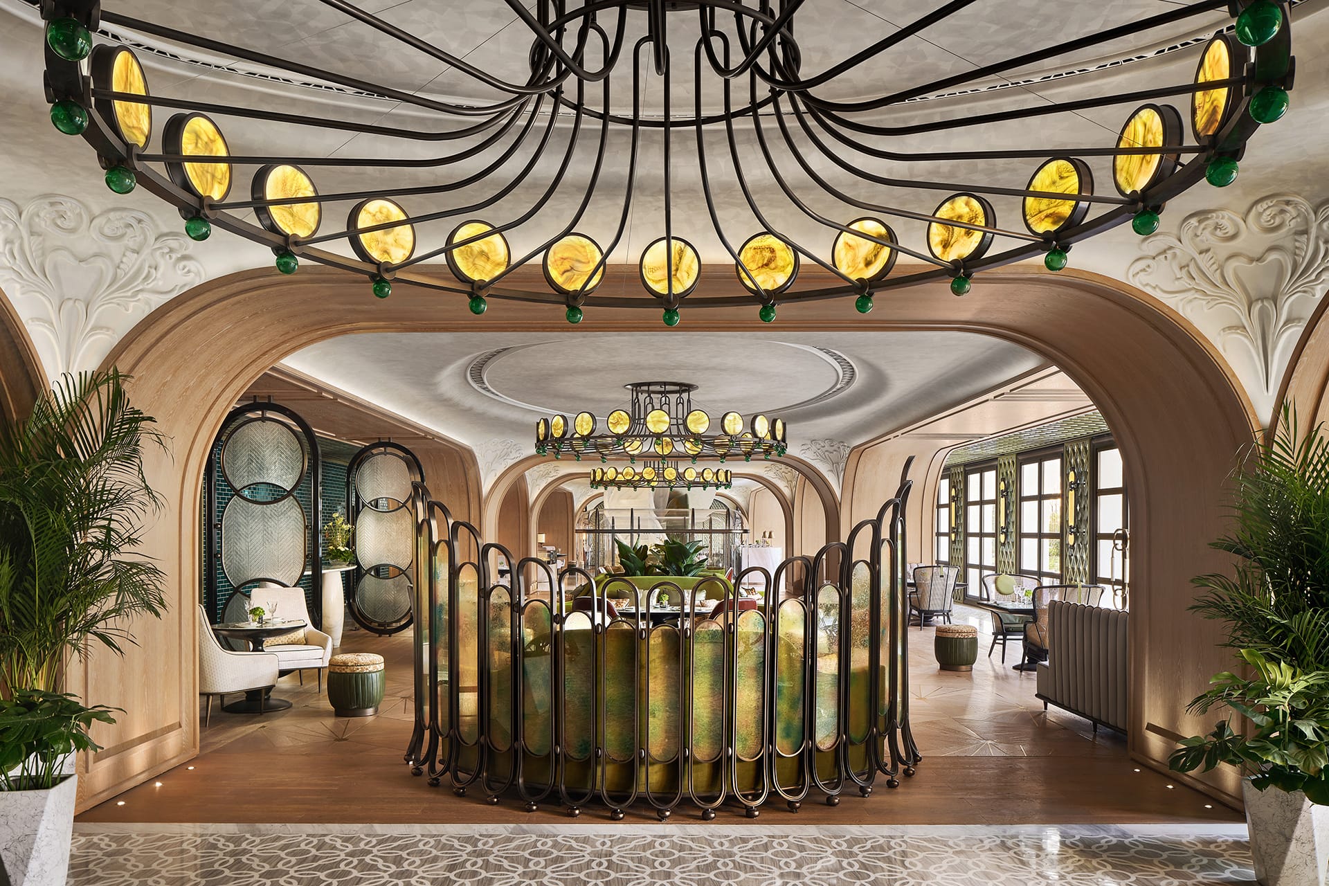 Art Deco room with ornate chandelier, green and gold decor, arches, and patterned floor tiles, surrounded by lush plants.