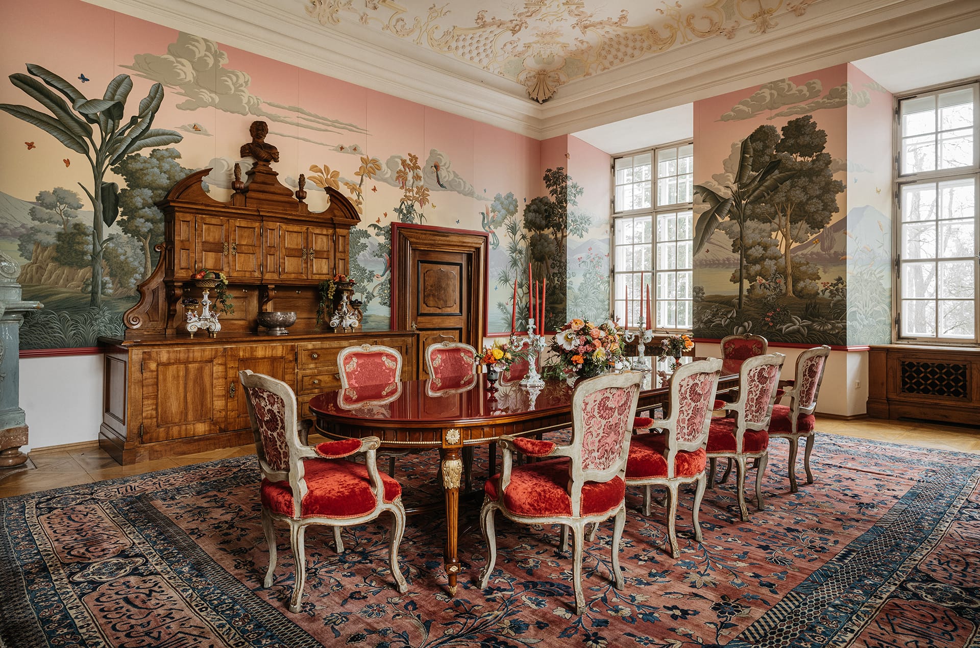 Elegant dining room with ornate pink wallpaper, red upholstered chairs, wooden furniture, and floral centerpieces on a long table.
