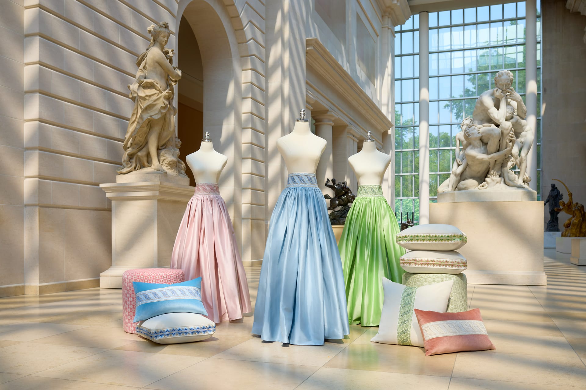 Three mannequins in pastel skirts displayed in a sunlit museum interior with decorative pillows nearby.