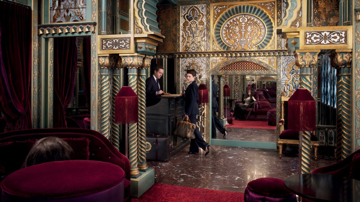 Ornate hotel lobby with people at a reception desk, featuring rich red velvet furnishings and intricate gold and green decor.