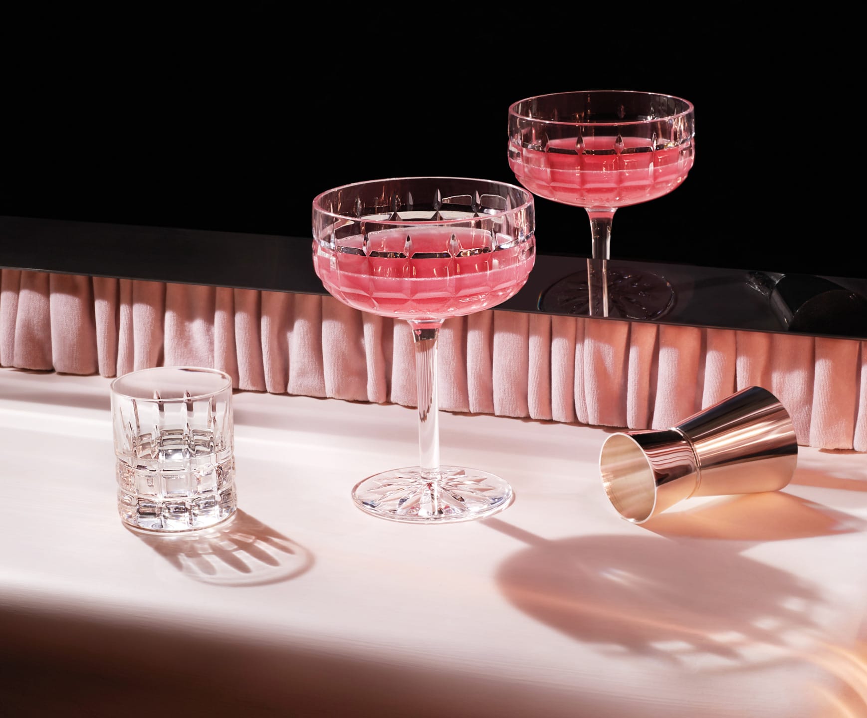 Two crystal cocktail glasses filled with pink drink, next to an empty glass and a metal shaker on a bar counter.
