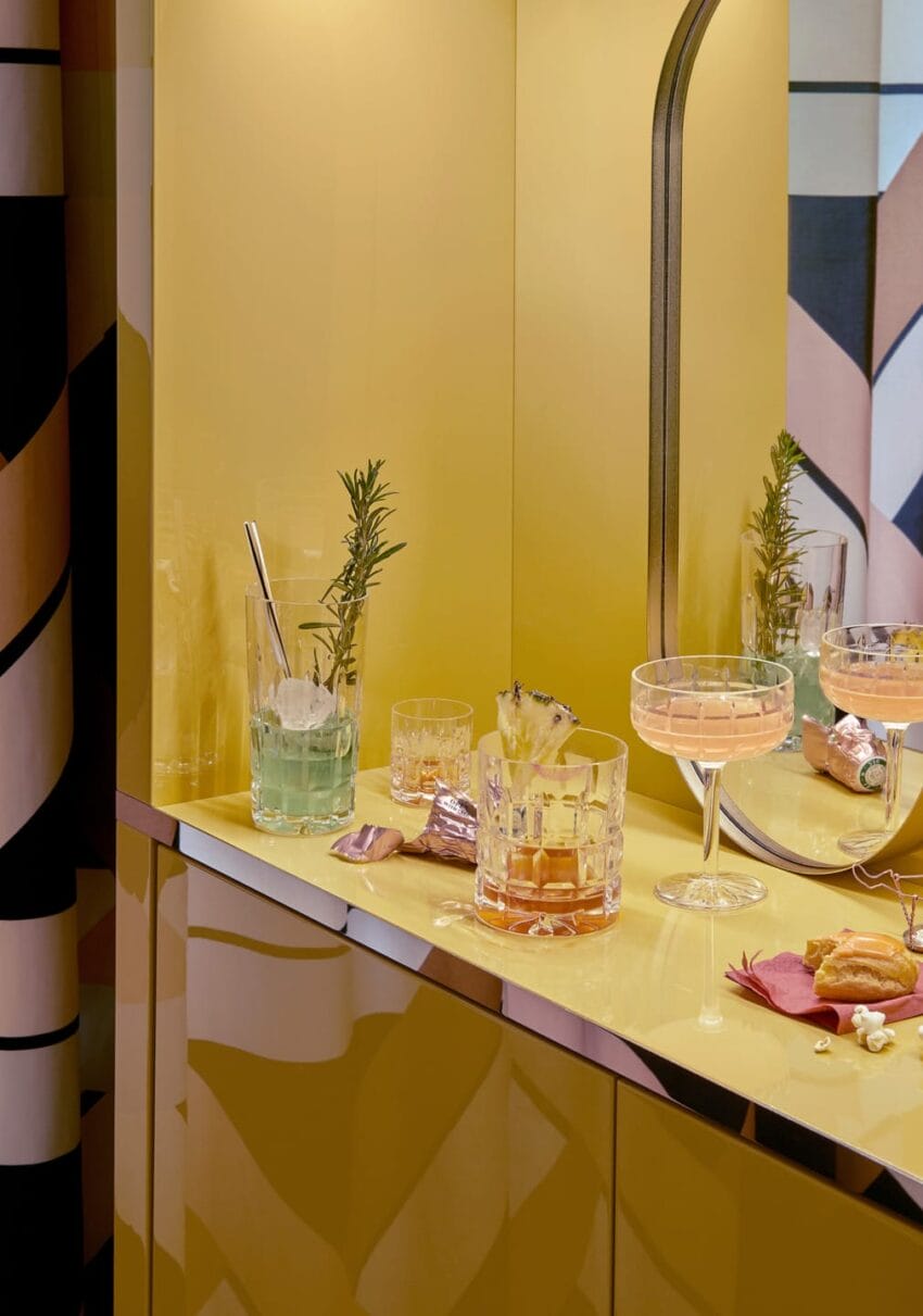 Cocktail glasses with drinks and garnishes on a yellow countertop with a mirrored backsplash.