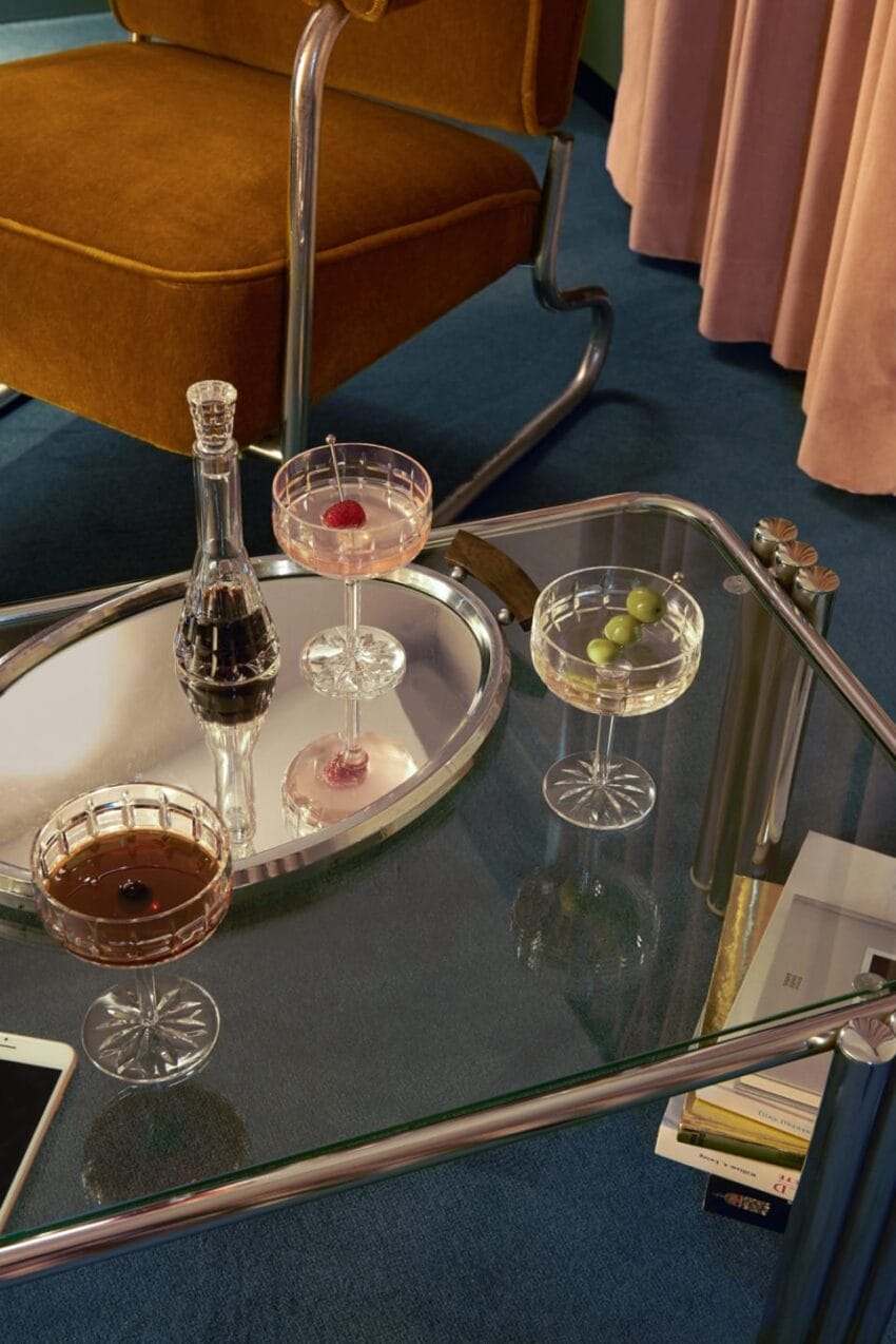 Modern glass table with elegant crystal glasses, wine decanter, and leather chair on a blue carpeted floor.