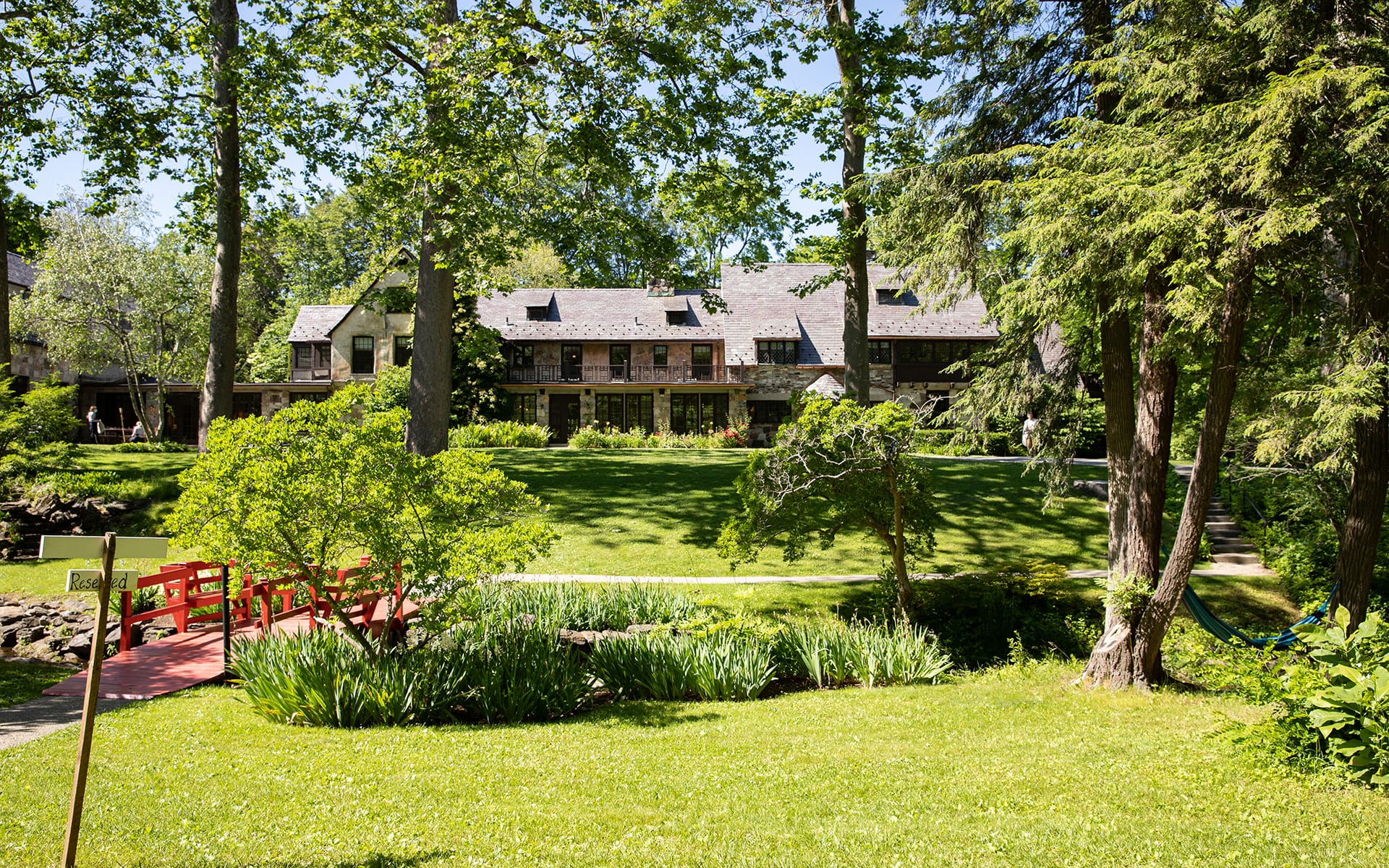 Scenic view of a large, rustic house surrounded by trees and a green lawn with a red footbridge in the foreground.