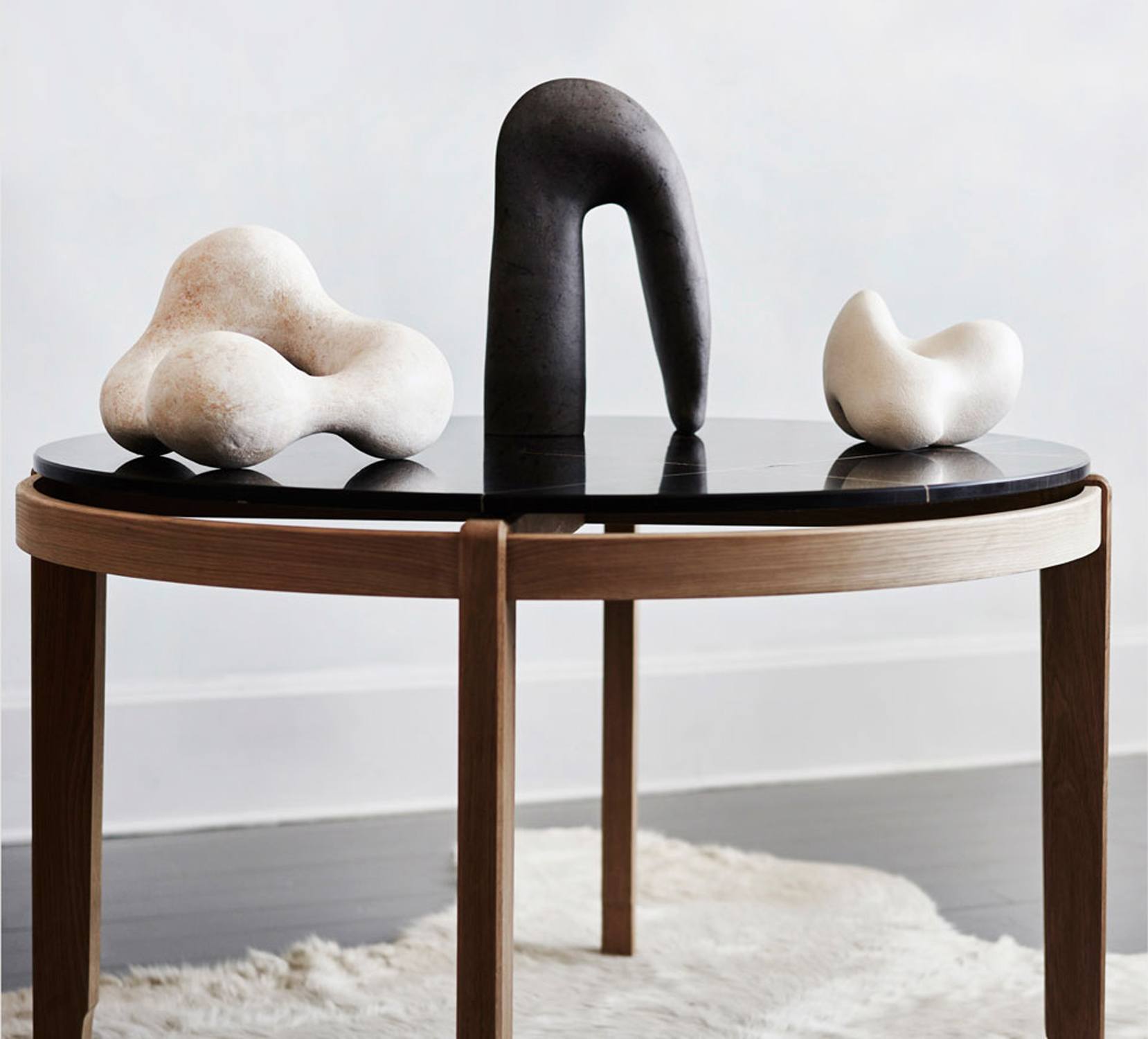 Modern abstract sculptures on a round wooden table with a black top, displayed on a white fur rug in a minimalist room
