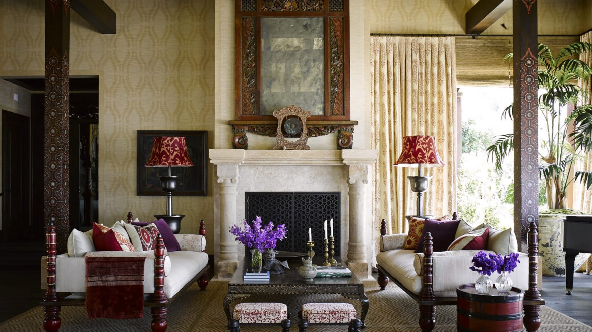Elegant living room with chandeliers, ornate fireplace, decorative rug, two sofas, floral cushions, and red lamps.
