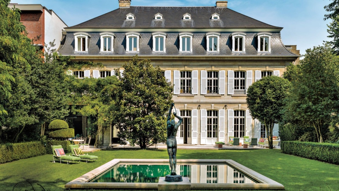 Elegant mansion with symmetrical windows and a manicured garden featuring a central sculpture and rectangular pool.