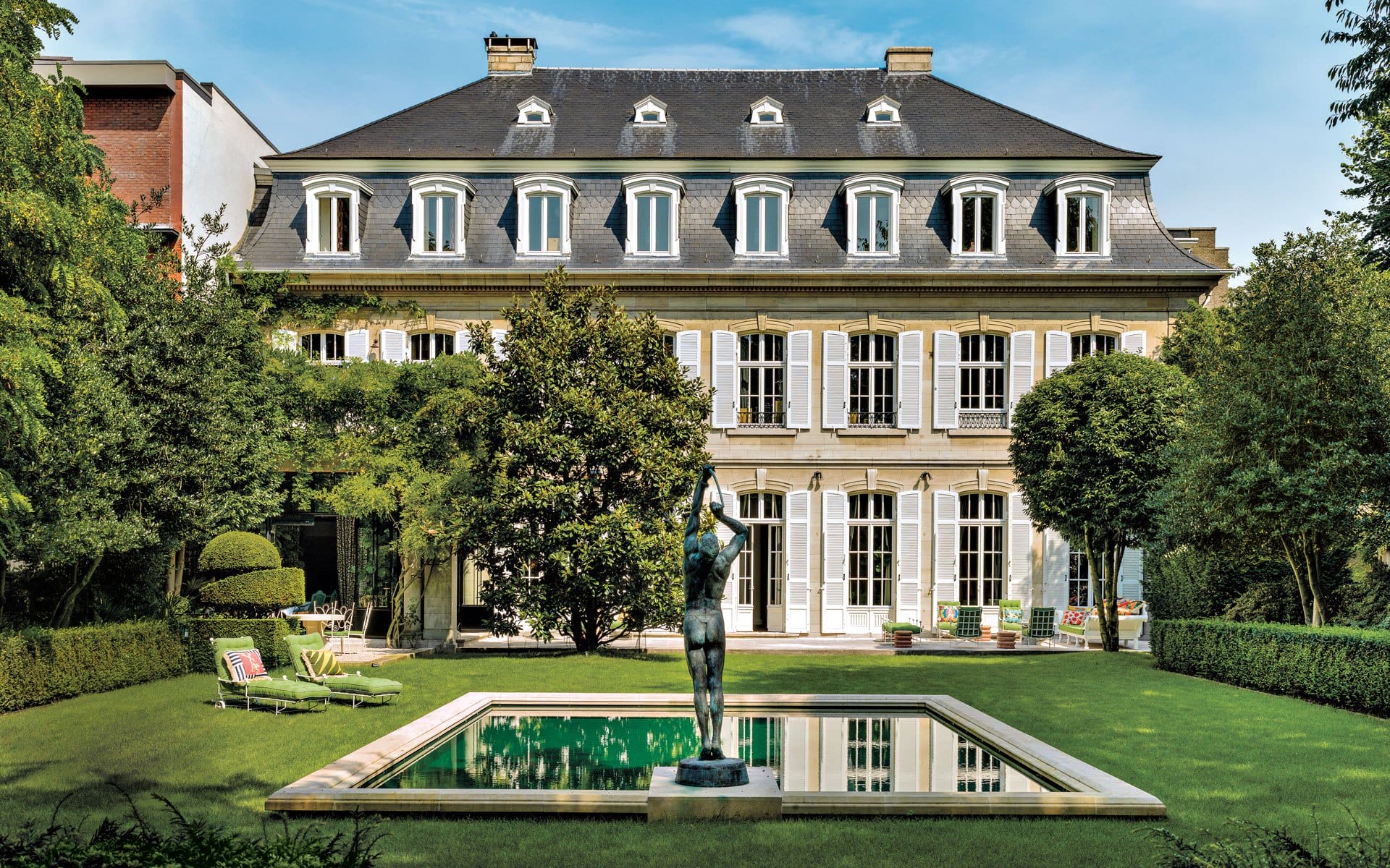 Elegant mansion with symmetrical windows and a manicured garden featuring a central sculpture and rectangular pool.