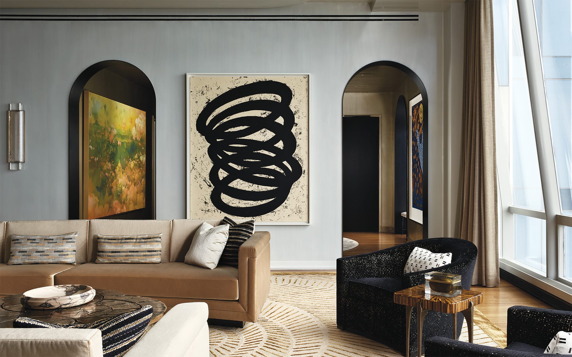 Modern living room with abstract art, arched doorways, beige sofa, black chair, and large windows with curtains.