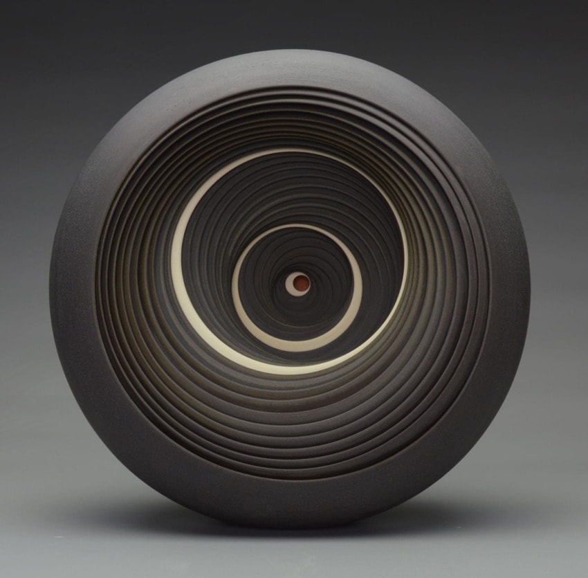 Abstract concentric circular sculpture with layered design in shades of black, white, and gray against a dark background.