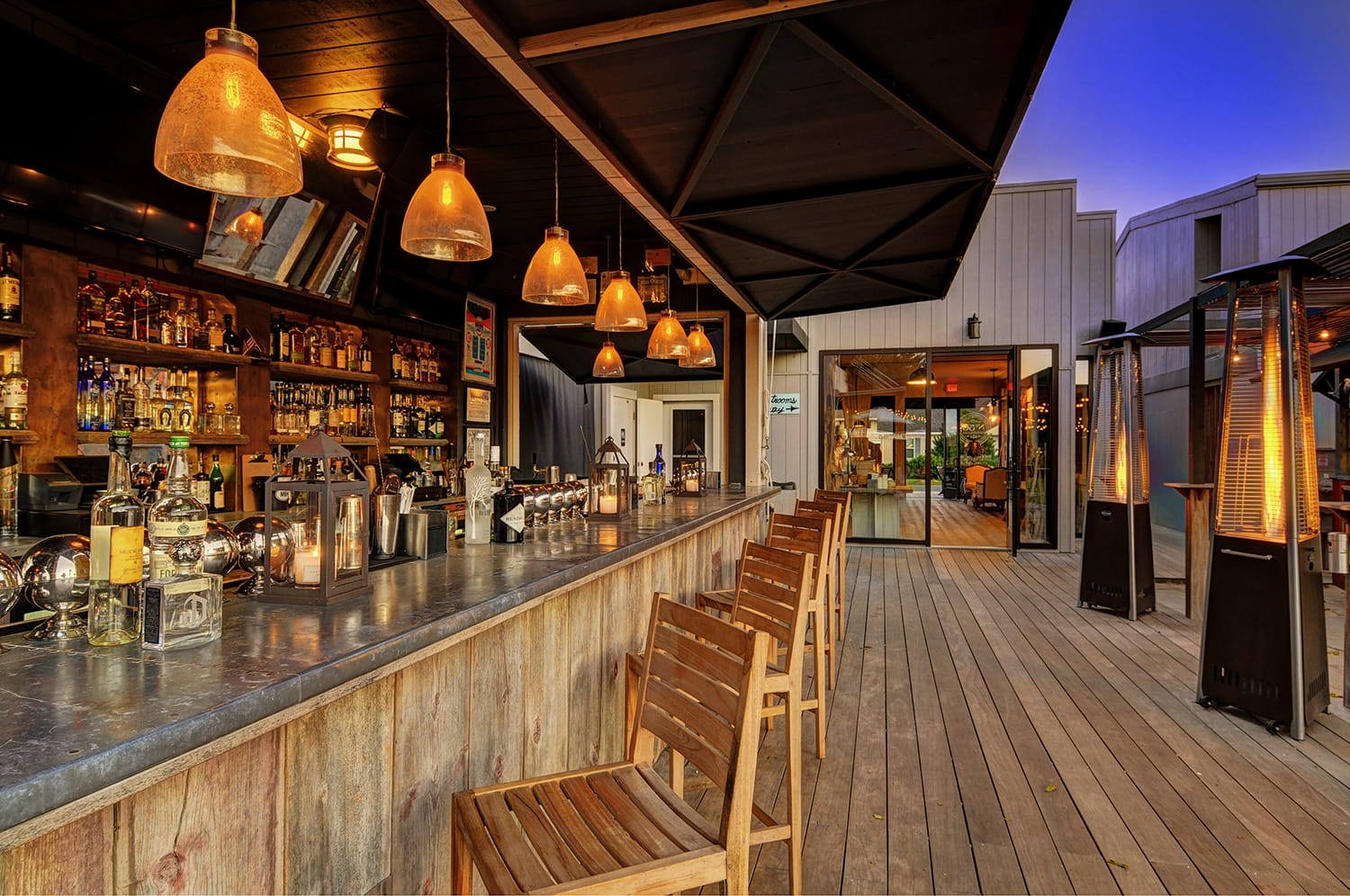 Outdoor bar with wooden chairs, hanging lights, and a variety of bottles on shelves, creating a warm, inviting atmosphere.