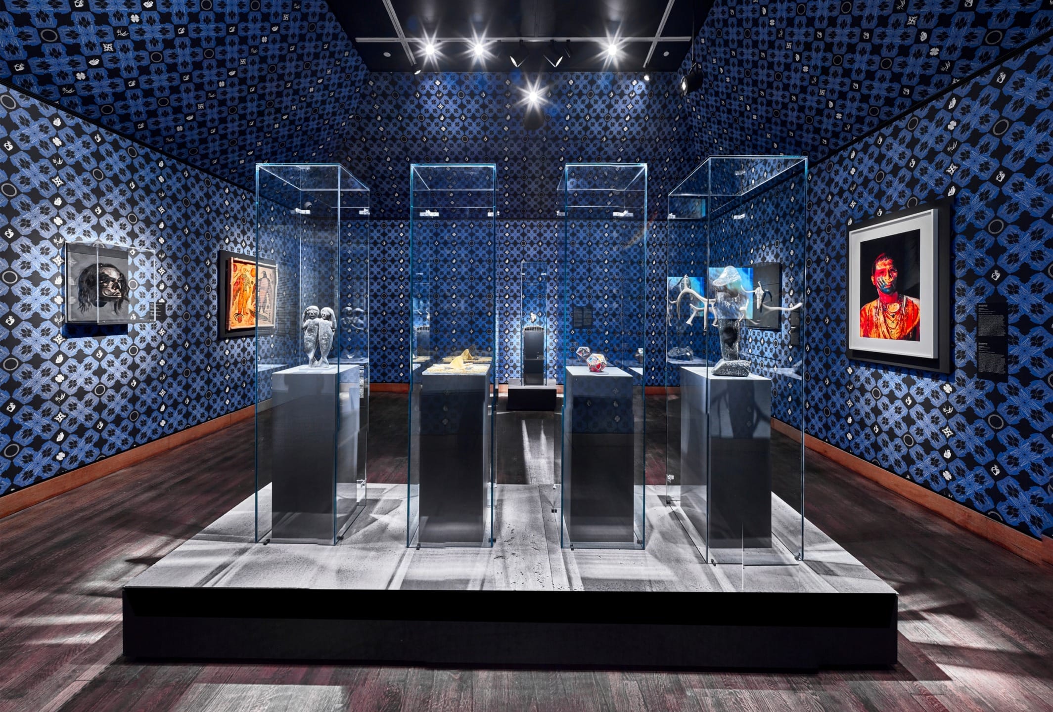 Museum exhibit room with blue patterned walls displaying art pieces in glass cases and framed artwork on the walls.