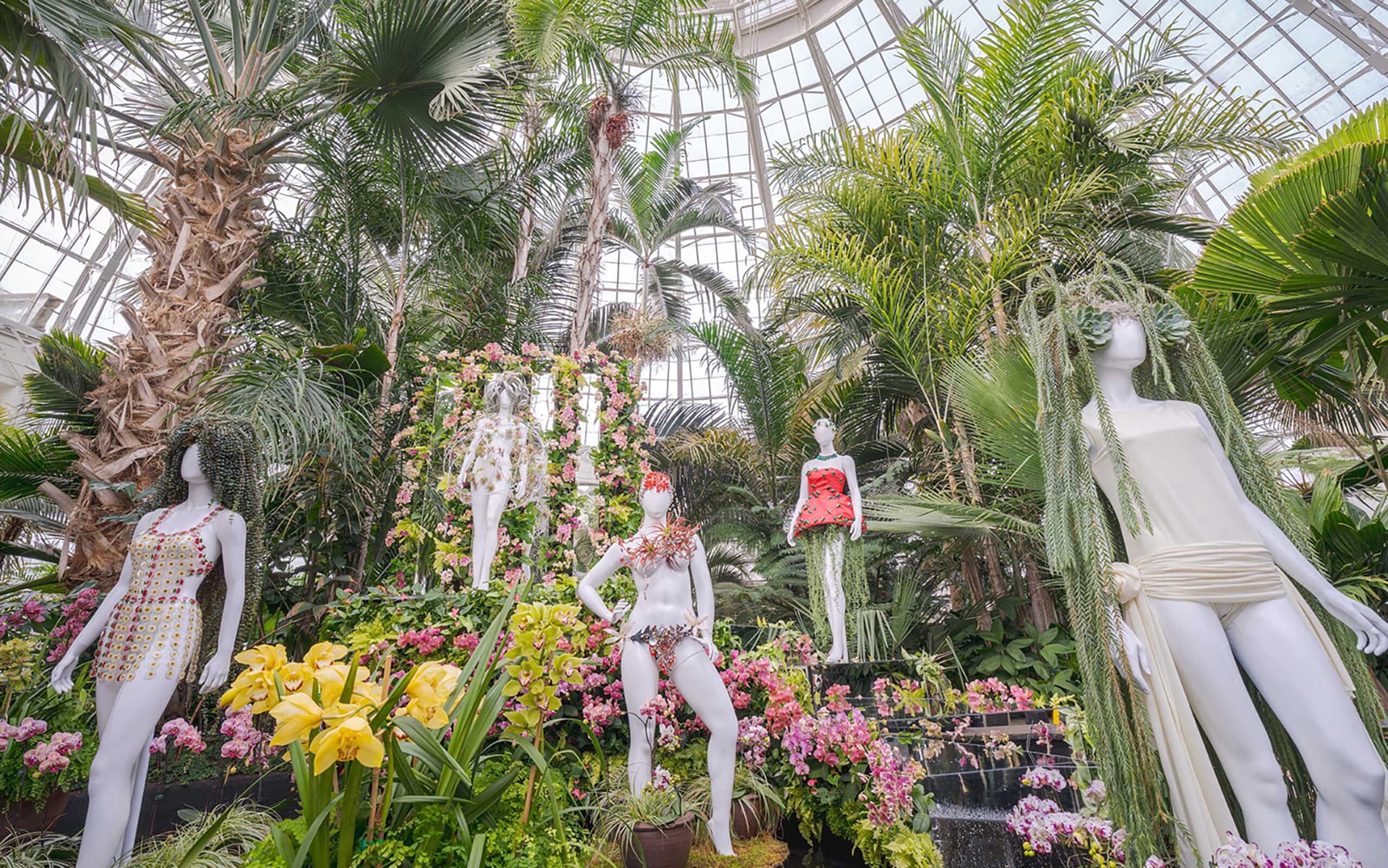 Mannequins adorned with floral dresses among lush greenery in a grand greenhouse setting.