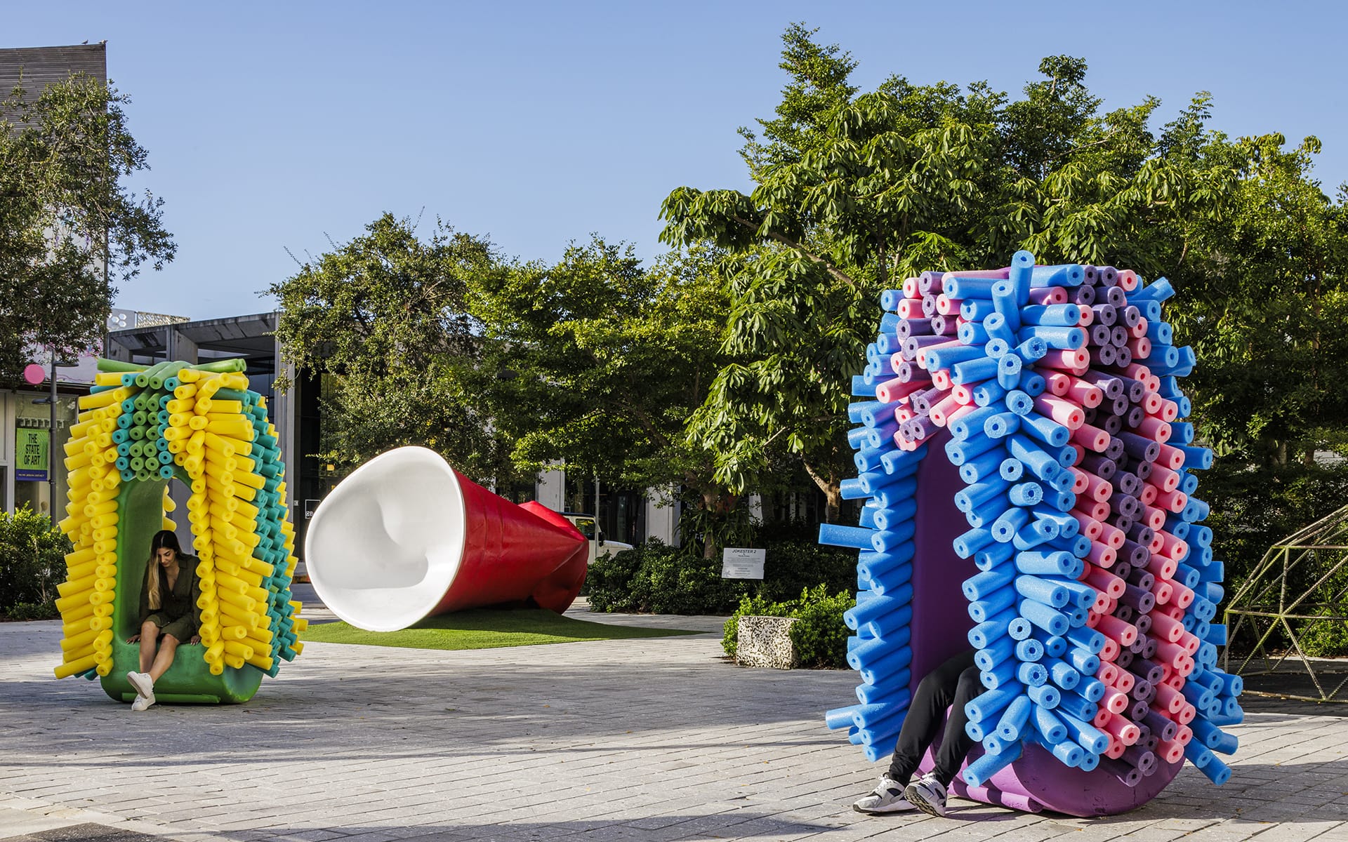 Colorful noodle art installations in a public park with trees and clear blue sky.