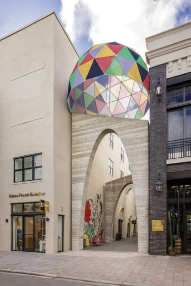 Colorful geometric dome atop modern building with archway, mural, and adjacent shops, under a partly cloudy sky.
