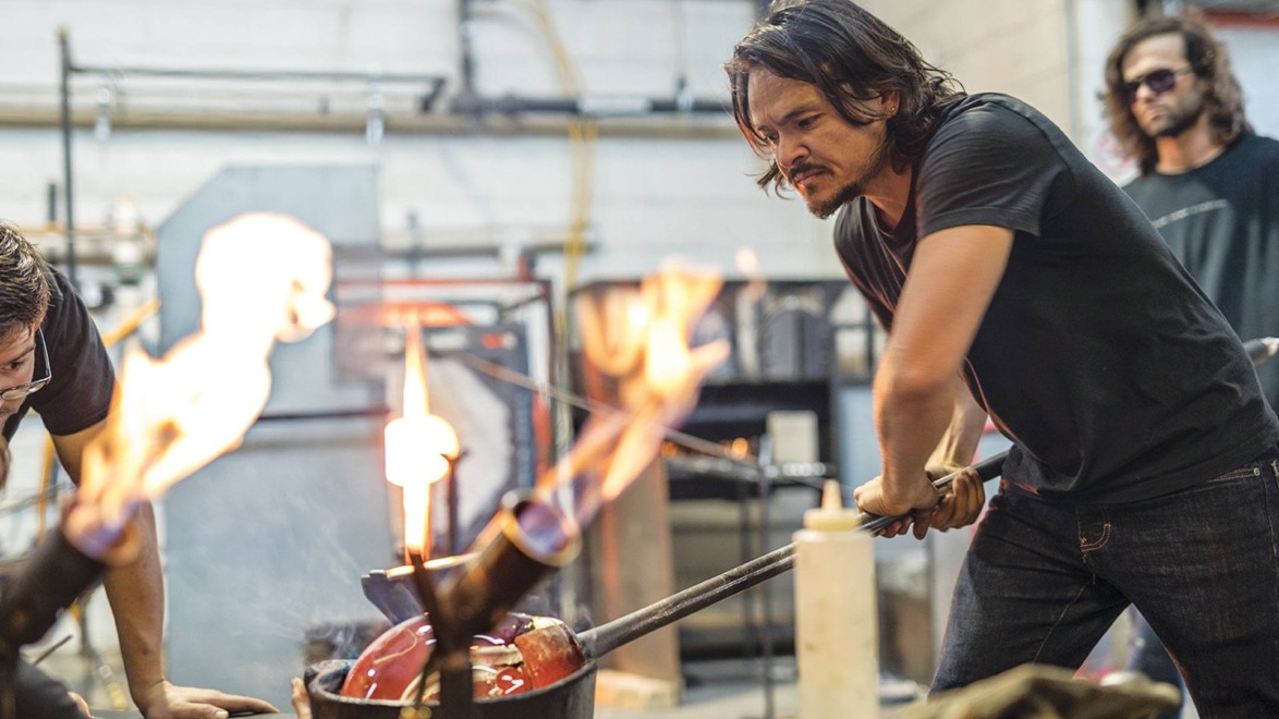 Glassblower shaping molten glass with intense focus in an industrial studio setting, surrounded by flames and equipment.