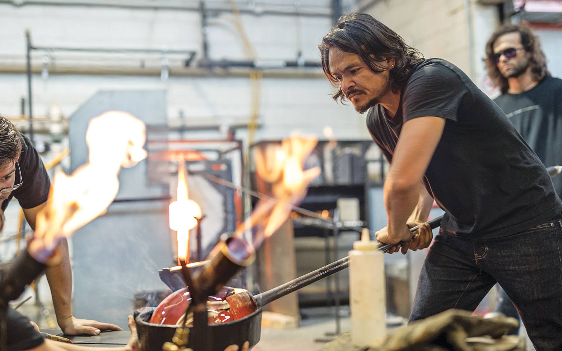 Glassblower shaping molten glass with intense focus in an industrial studio setting, surrounded by flames and equipment.
