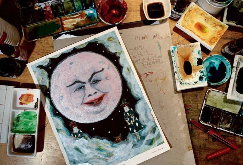Painting of a whimsical moon face surrounded by various paints and art supplies on a wooden table.
