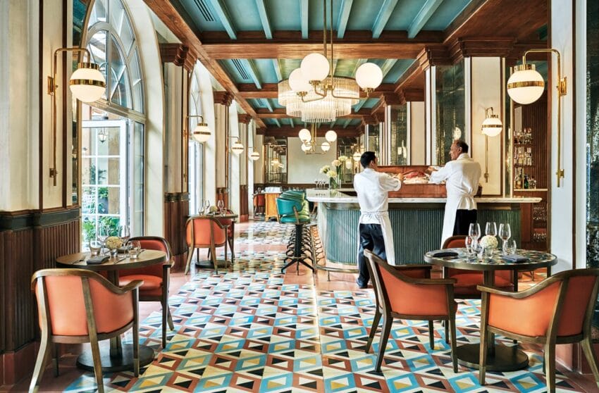 Elegant restaurant interior with colorful geometric tiled floor, round tables, leather chairs, and two chefs behind a counter.