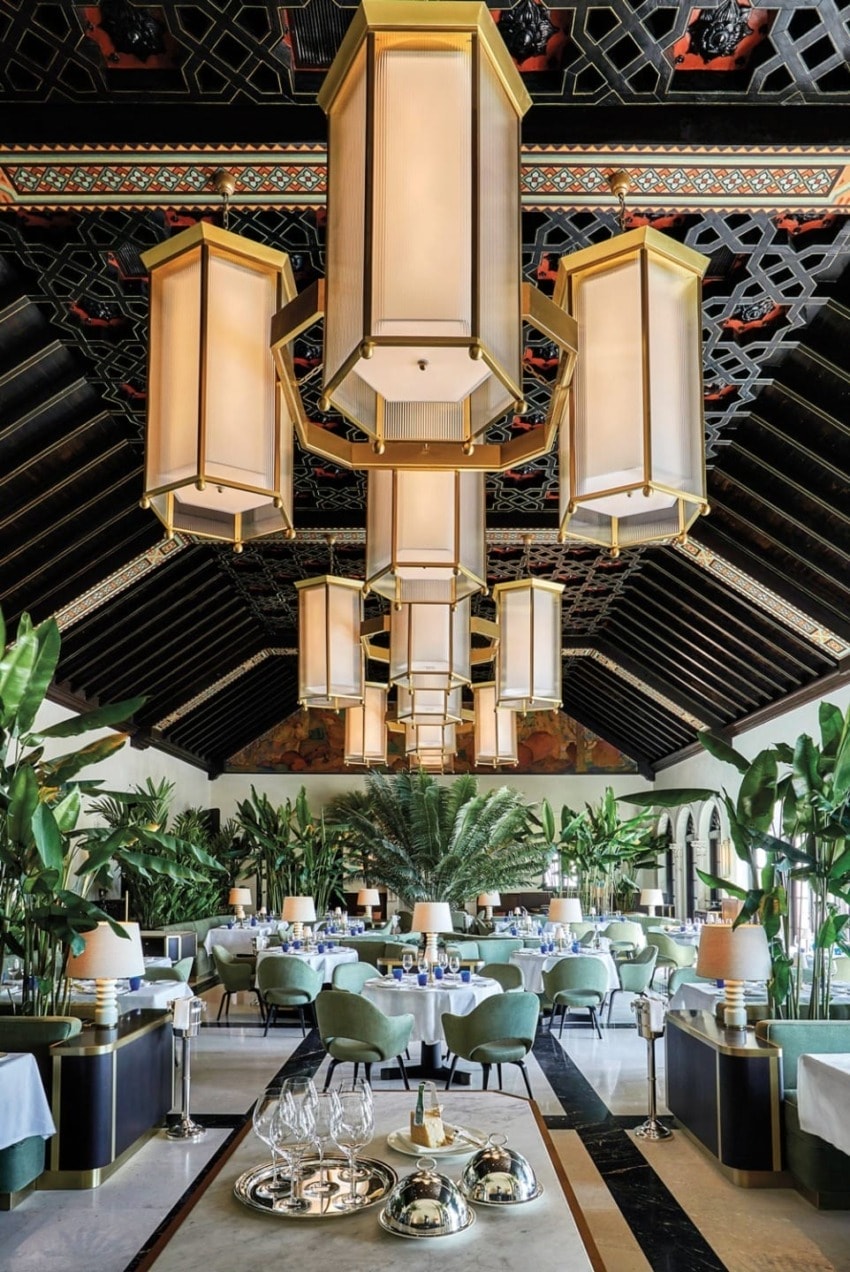 Luxurious restaurant with elegant chandeliers, lush green plants, and neatly set tables in a grand dining area.
