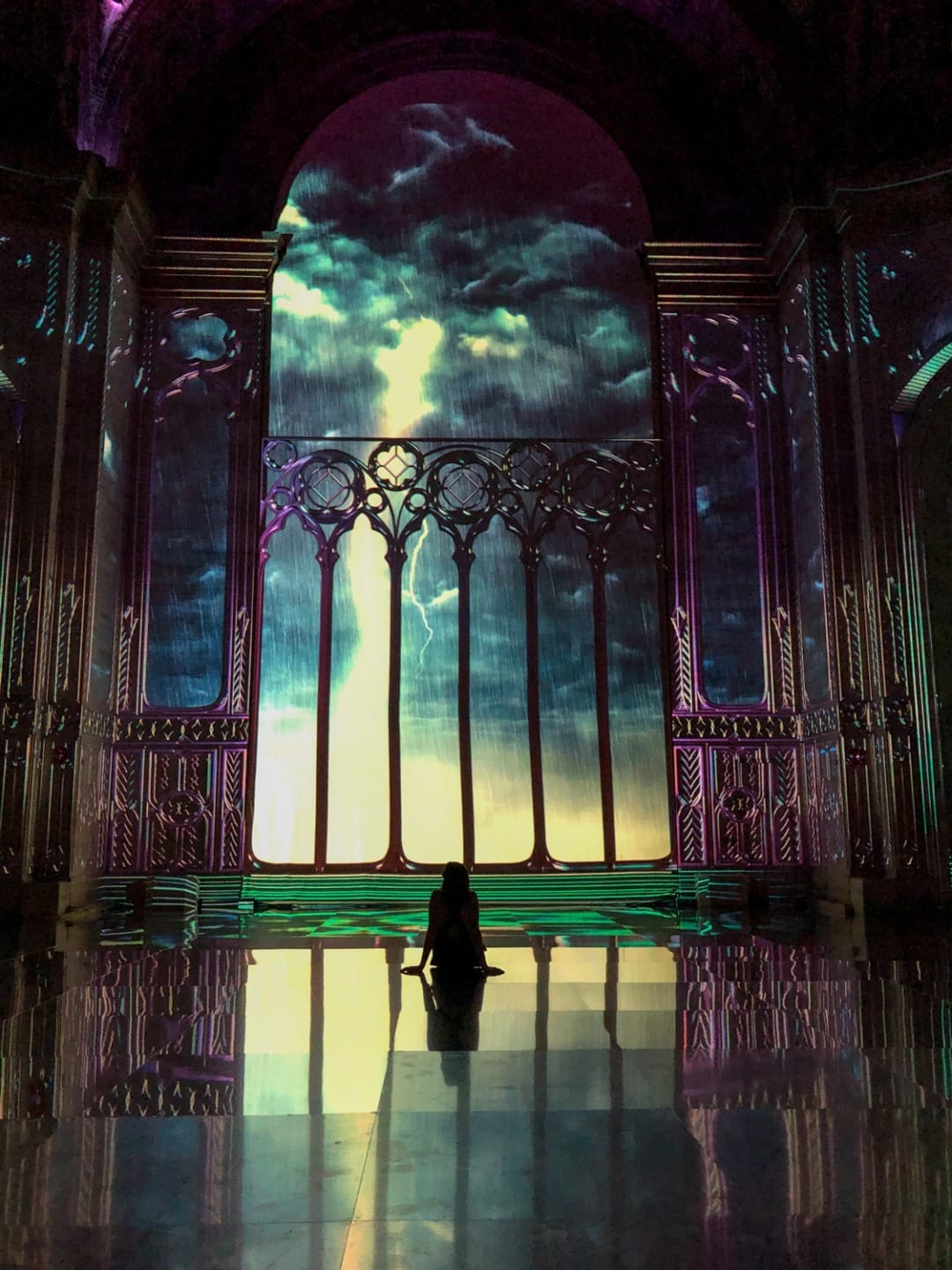 Silhouette of a person sitting before an ornate, illuminated window in a grand hall with a cosmic sky projection.