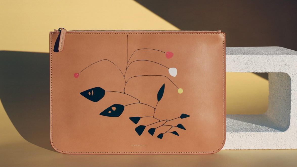 Brown leather clutch with abstract design, showcasing colorful geometric shapes, set on beige and stone backdrop.