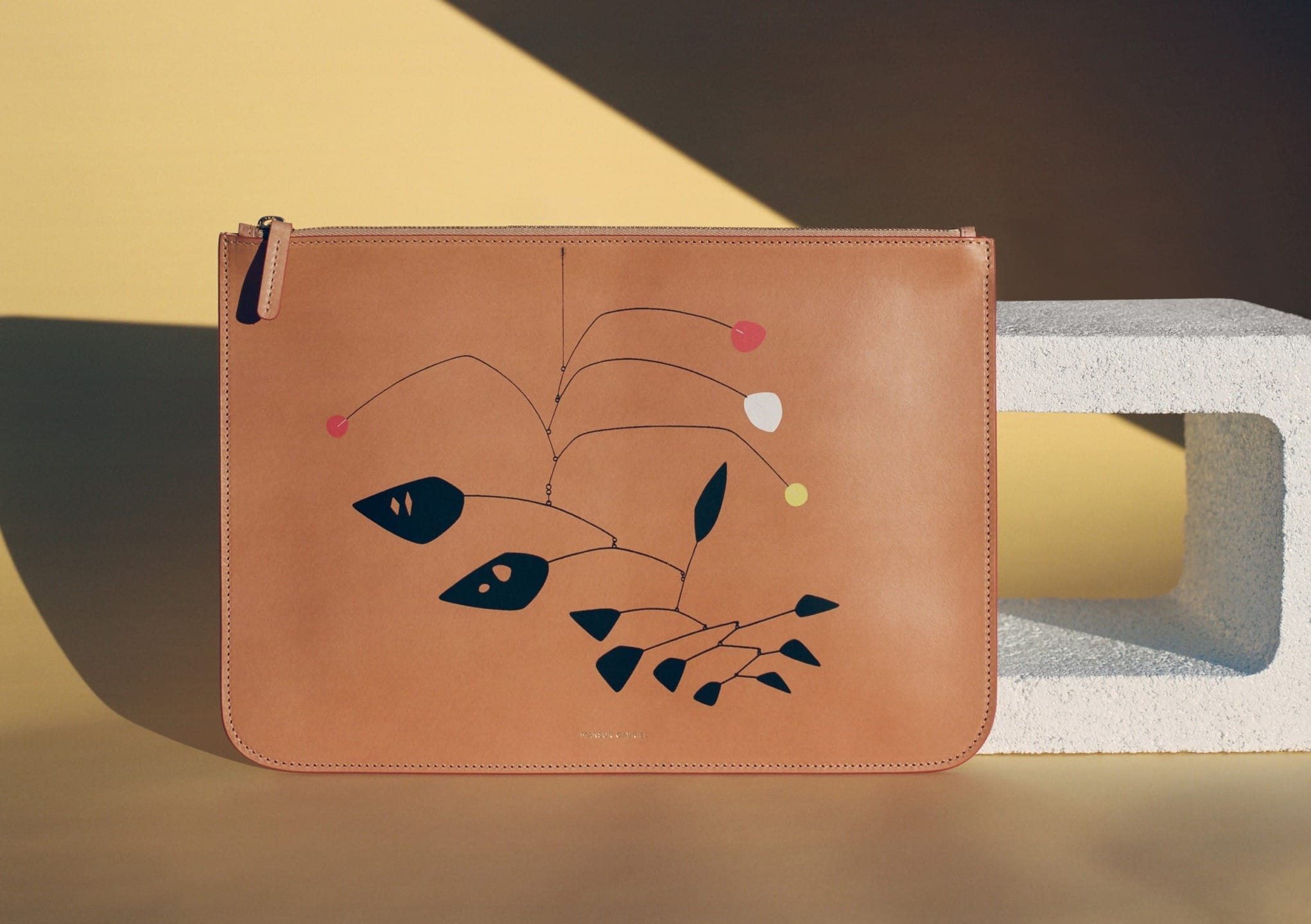 Brown leather clutch with abstract design, showcasing colorful geometric shapes, set on beige and stone backdrop.