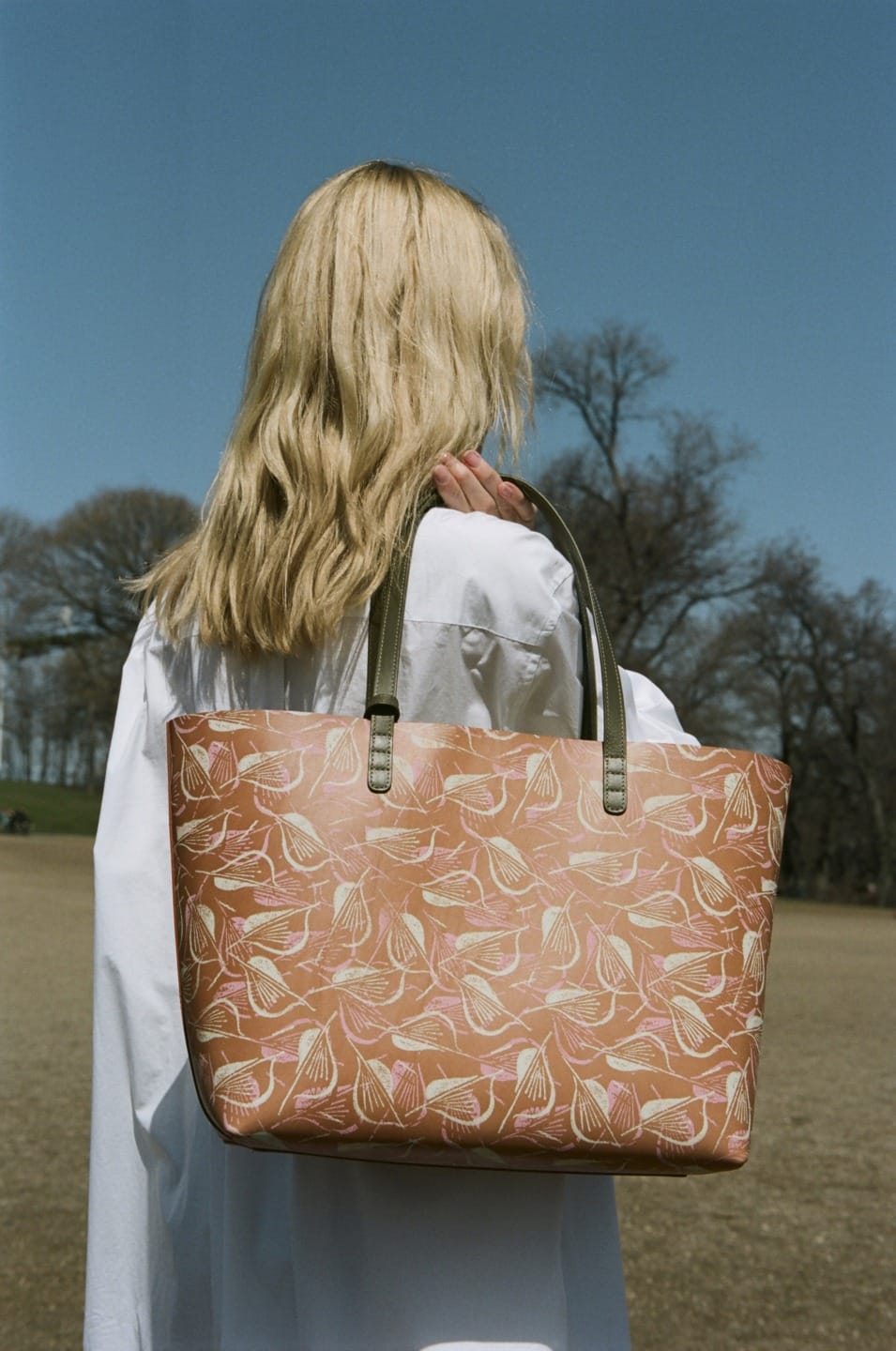 Woman with long hair holding a patterned tote bag outdoors with trees in the background.