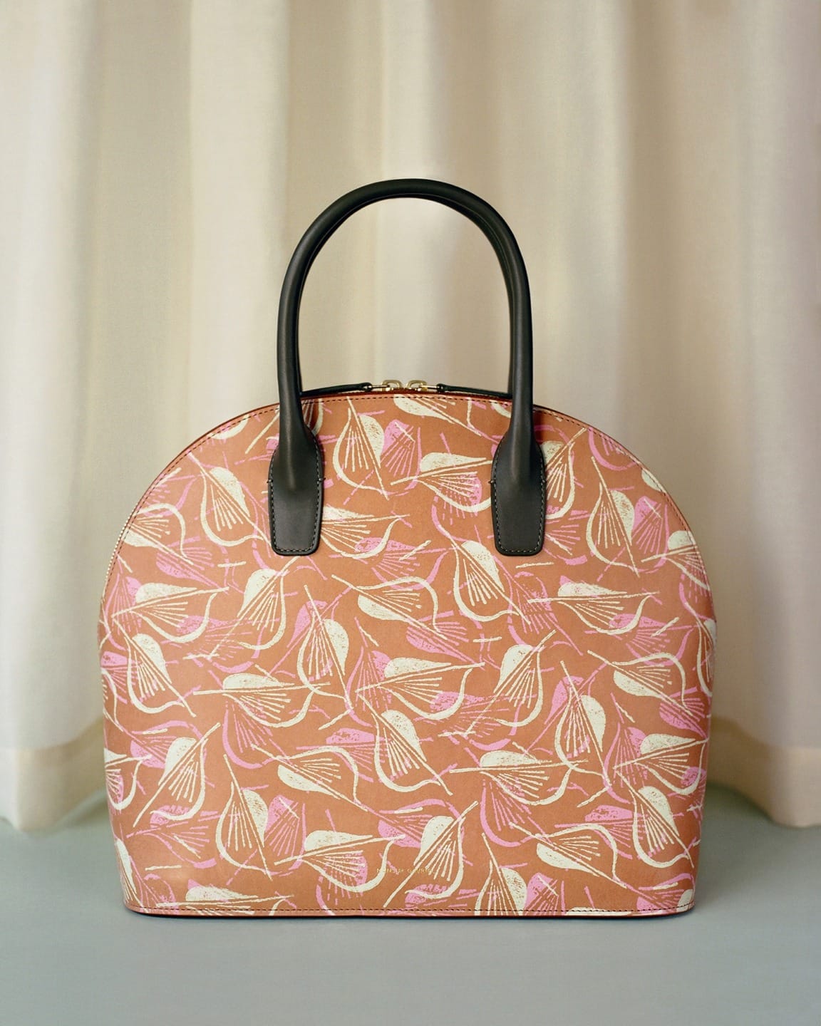 Patterned handbag with black handles, featuring pink and beige floral design, displayed in front of a cream curtain.