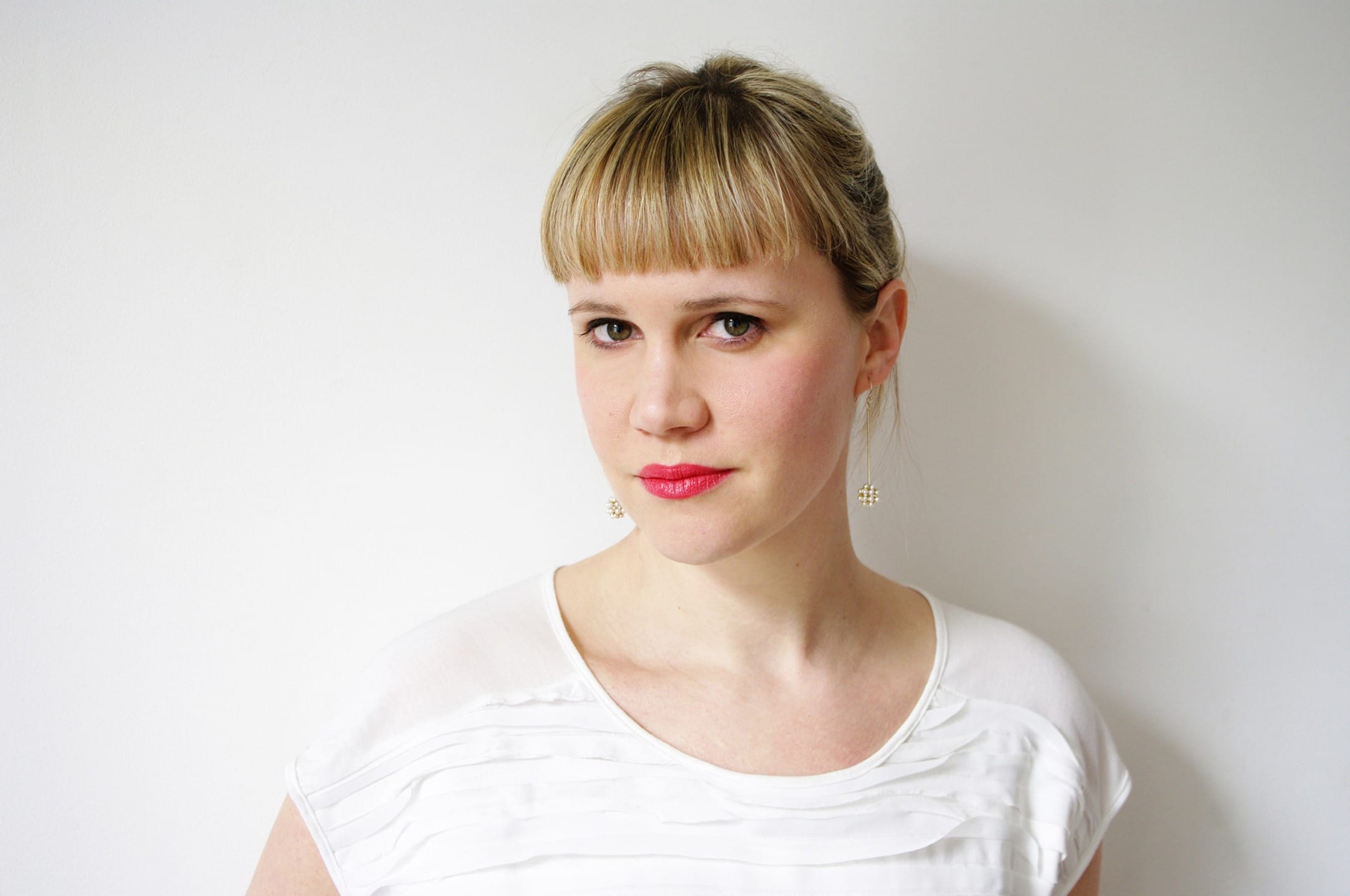 Person with blonde hair and bangs wearing a white shirt with a neutral expression, standing against a white background.