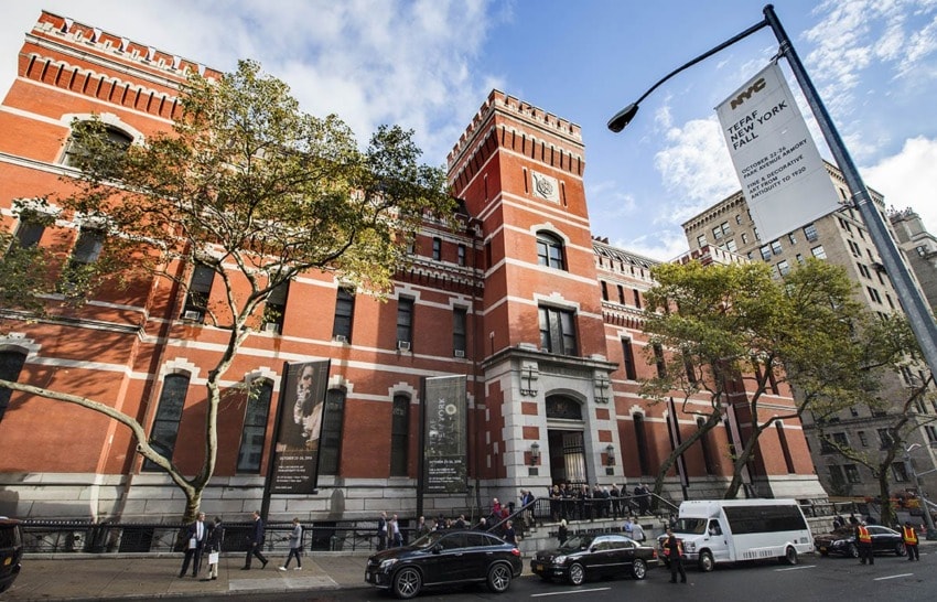 Red brick building with people and cars in front, trees lining the street, and a sign for Park Avenue Armory in New York City.