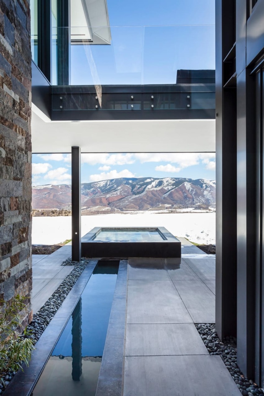 Modern architectural design featuring a glass walkway, mountain view, reflecting pool, and stone wall.