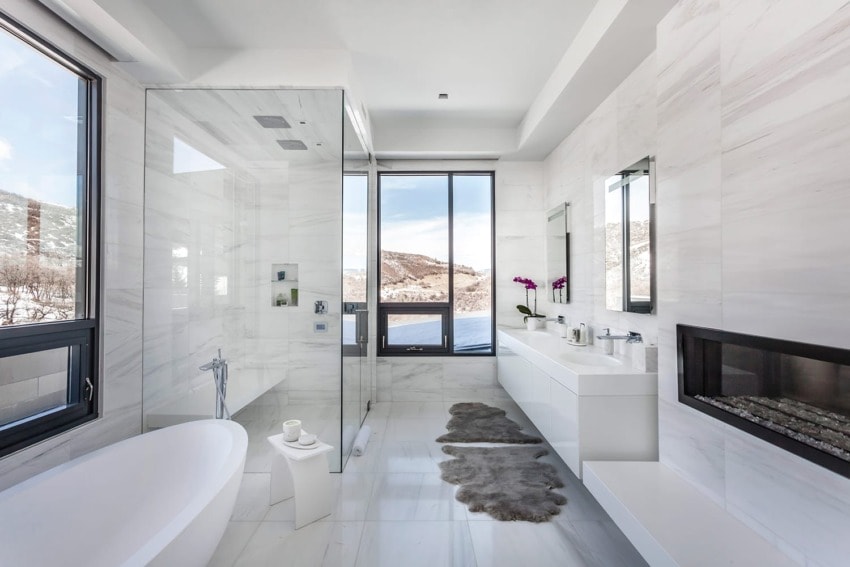 Modern bathroom with large windows, freestanding tub, walk-in shower, fireplace, and mountain view in the background.