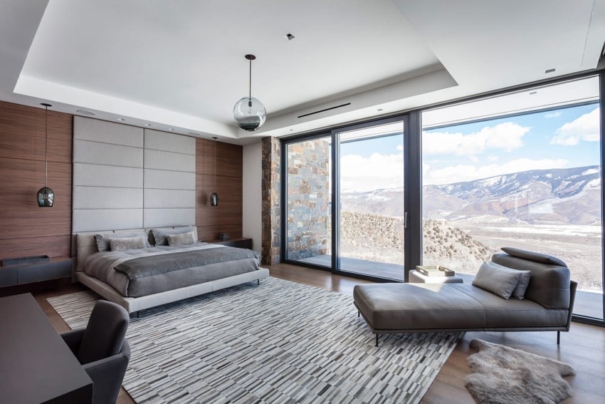 Modern bedroom with large windows, mountain view, gray bed, chaise lounge, and geometric rug on wooden floor.