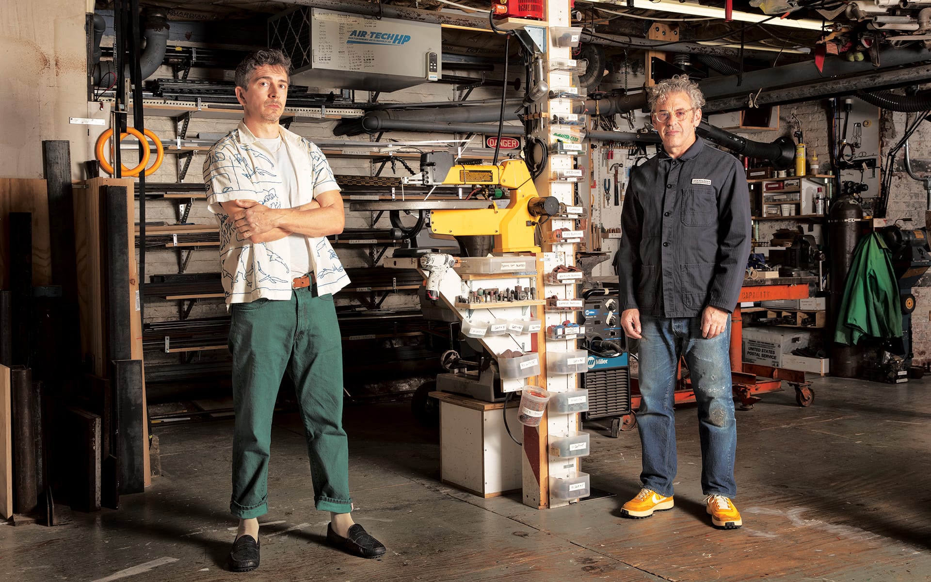 Two men stand in a workshop with various tools and equipment in the background. One is standing next to a tall stack of items.