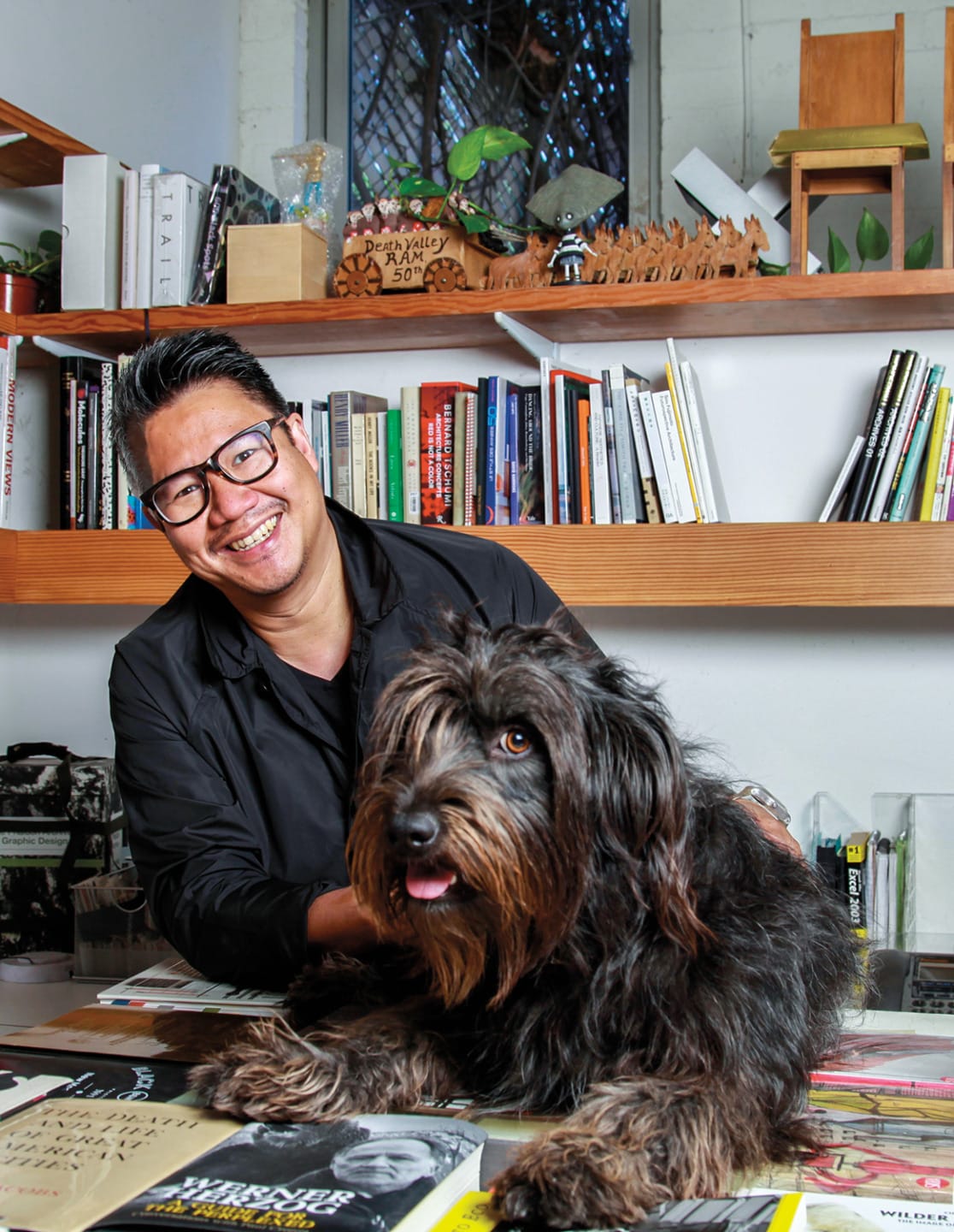 Man smiling while sitting at a desk with a fluffy black dog in a cozy room filled with books and plants.
