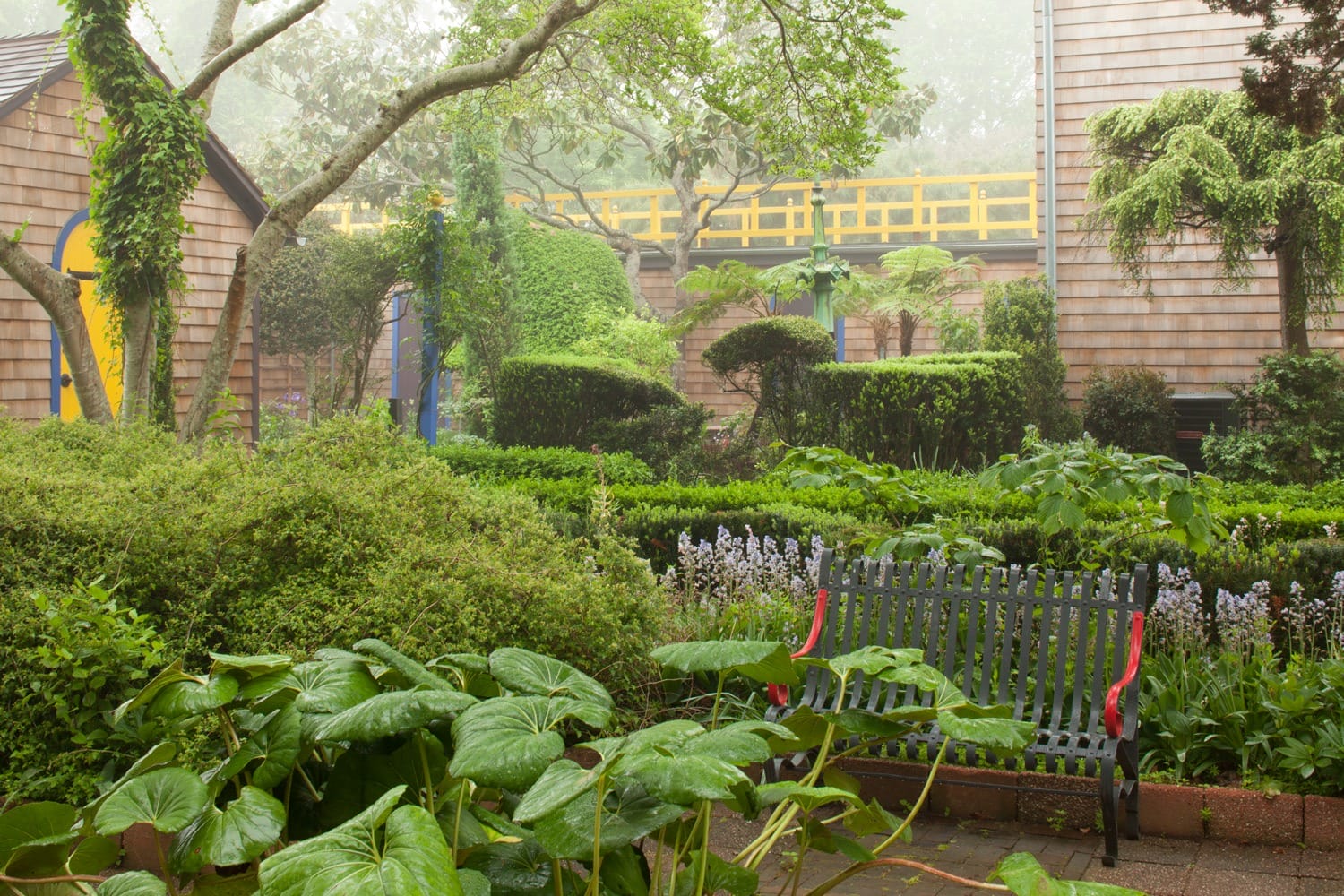 Serene garden with lush greenery, a wooden bench, and quaint wooden buildings surrounded by mist.