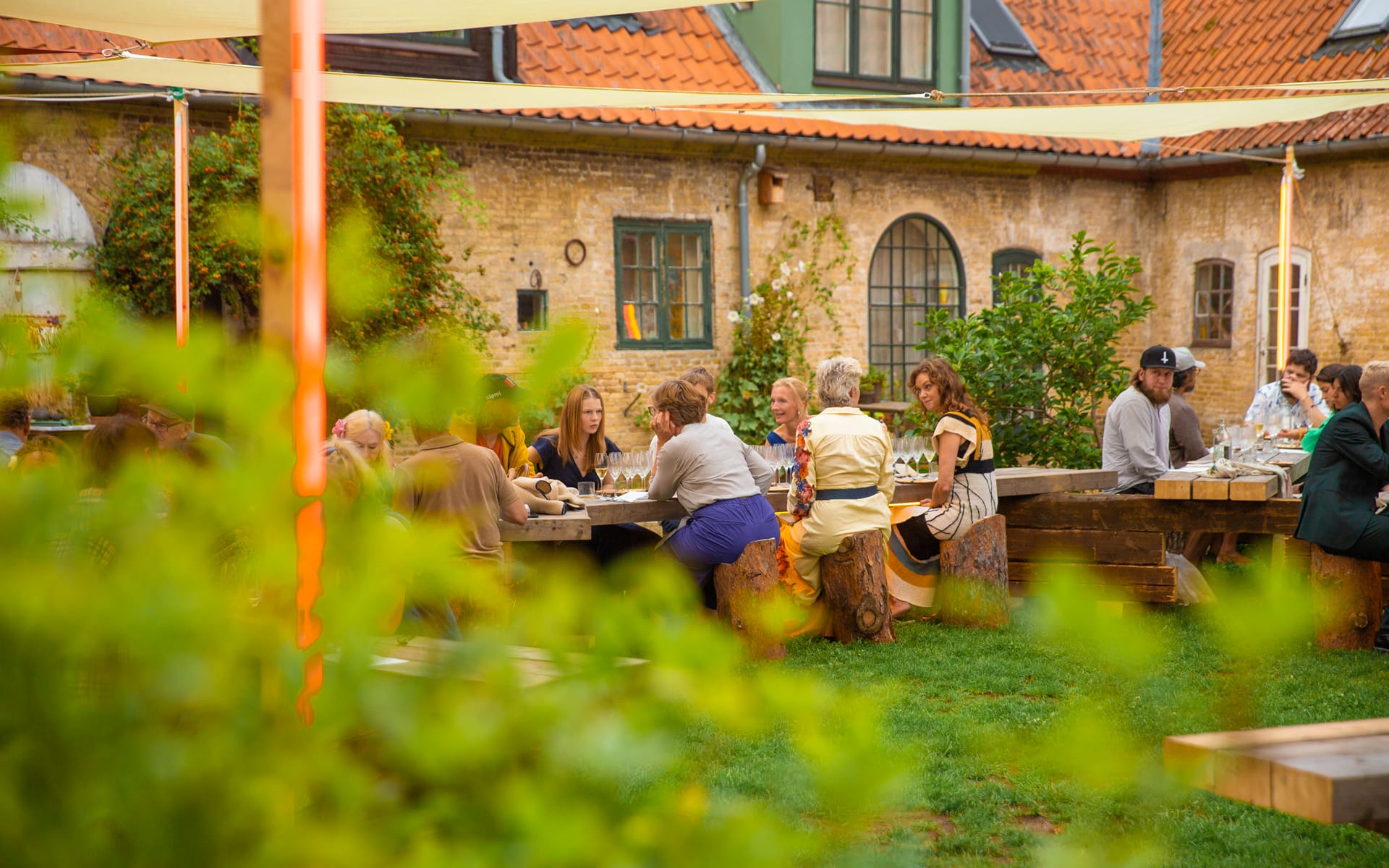 People enjoying a meal outdoors at wooden tables surrounded by greenery in a rustic courtyard setting.