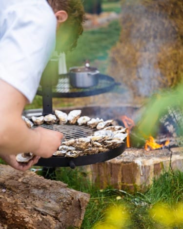 Person grilling oysters over an open flame outdoors with green foliage and hay bales in the background.