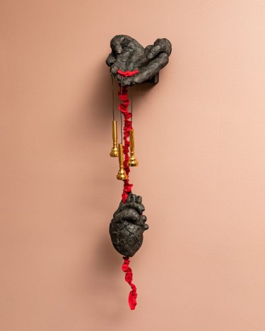 Sculpture of hands and heart with red ribbon and brass bells on peach wall background.