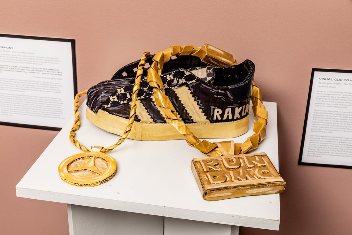 Cardboard sculpture of a sneaker with a gold chain and RUN DMC pendant on a white pedestal against a pink wall.