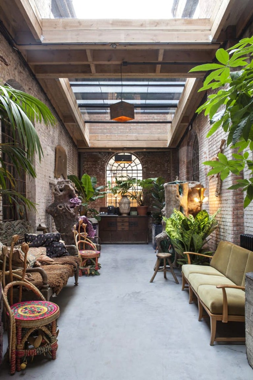 Eclectic indoor space with plants, wicker chairs, sofas, rustic brick walls, large windows, and a wooden ceiling.