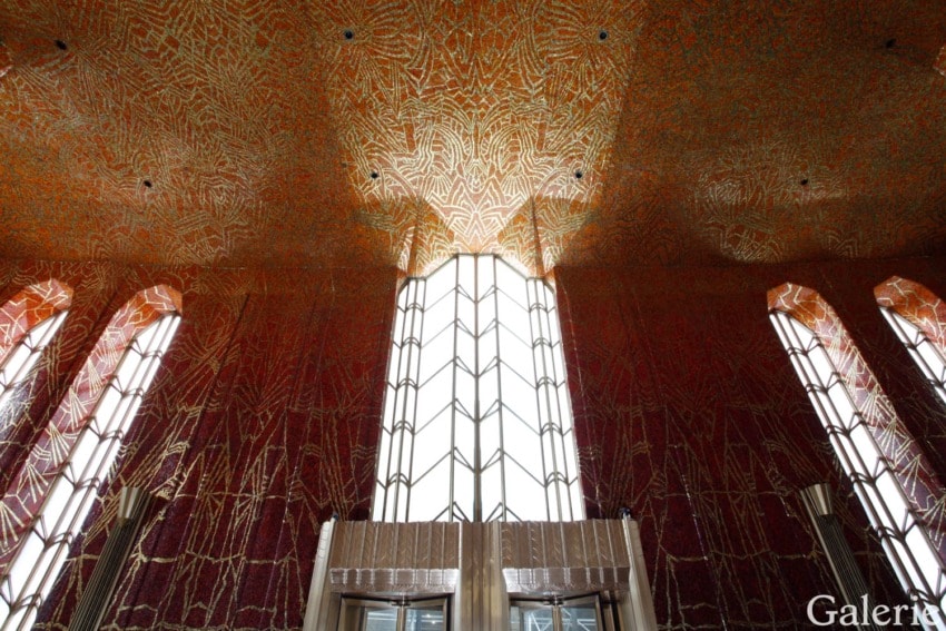 Interior of a building with tall, intricate stained glass windows and an ornate ceiling with geometric patterns.