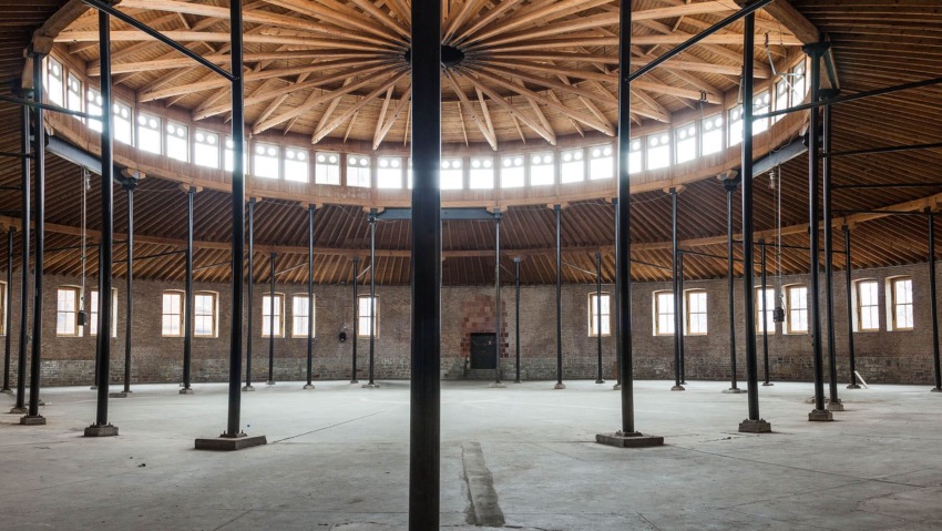 Interior of a large circular abandoned warehouse with a wooden ceiling and numerous metal support beams.